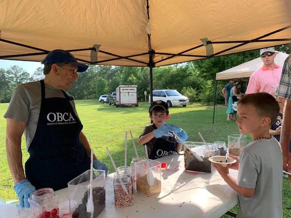 Man wearing a black apron with 'OBCA' logo and gloves standing at a table outdoors with children, showing various toppings in containers, under a canopy with other tents and people in the background.