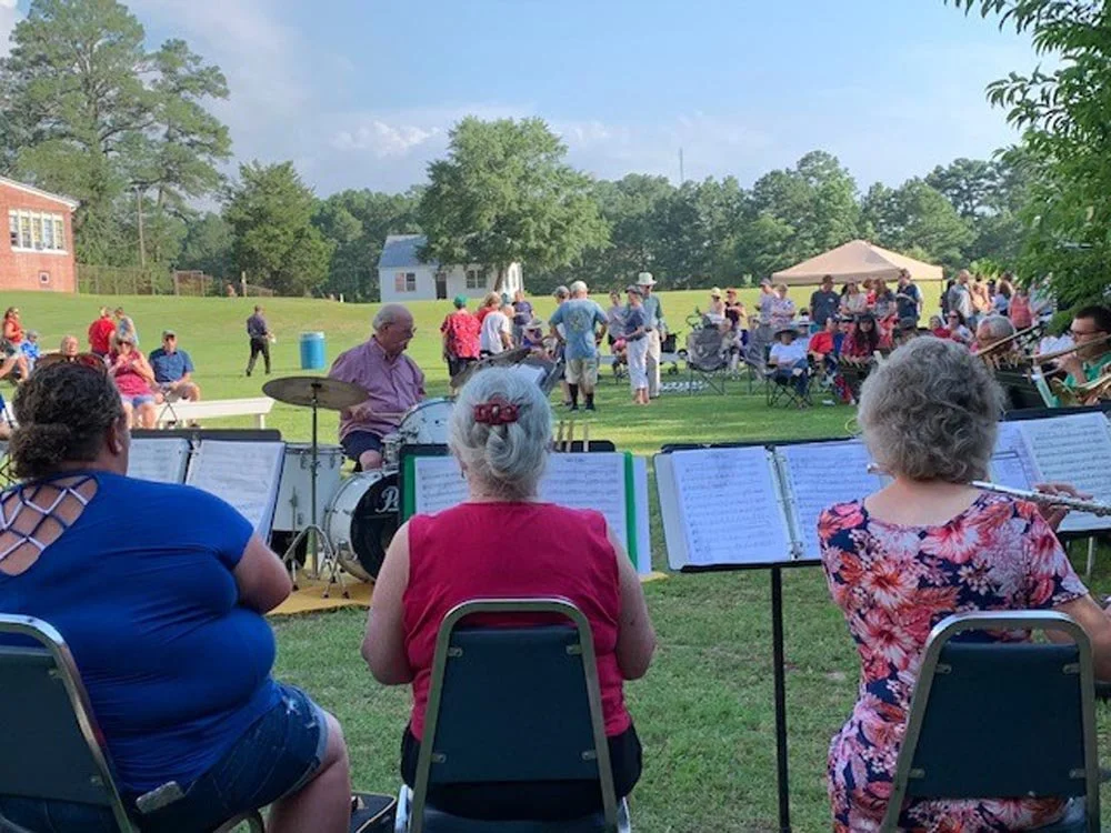 Outdoor community event with a band playing music, audience sitting and standing on a grassy field, and people enjoying a sunny day.