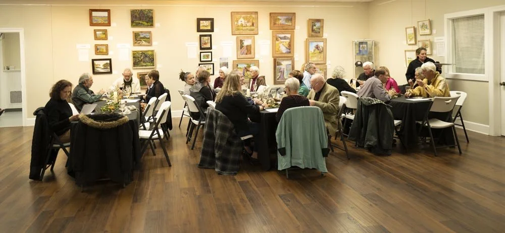 Group of elderly people dining together in an art gallery or community room with paintings on the wall
