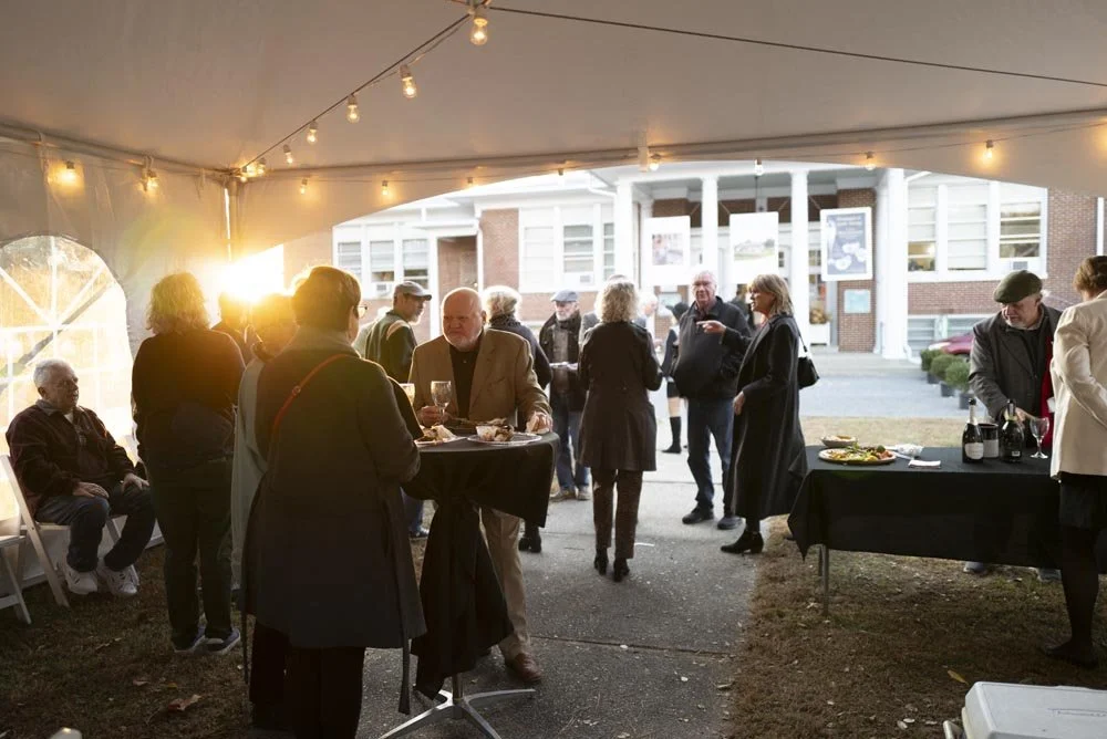 People socializing at an outdoor event under a canopy with string lights, with a building in the background and a table with food and drinks.