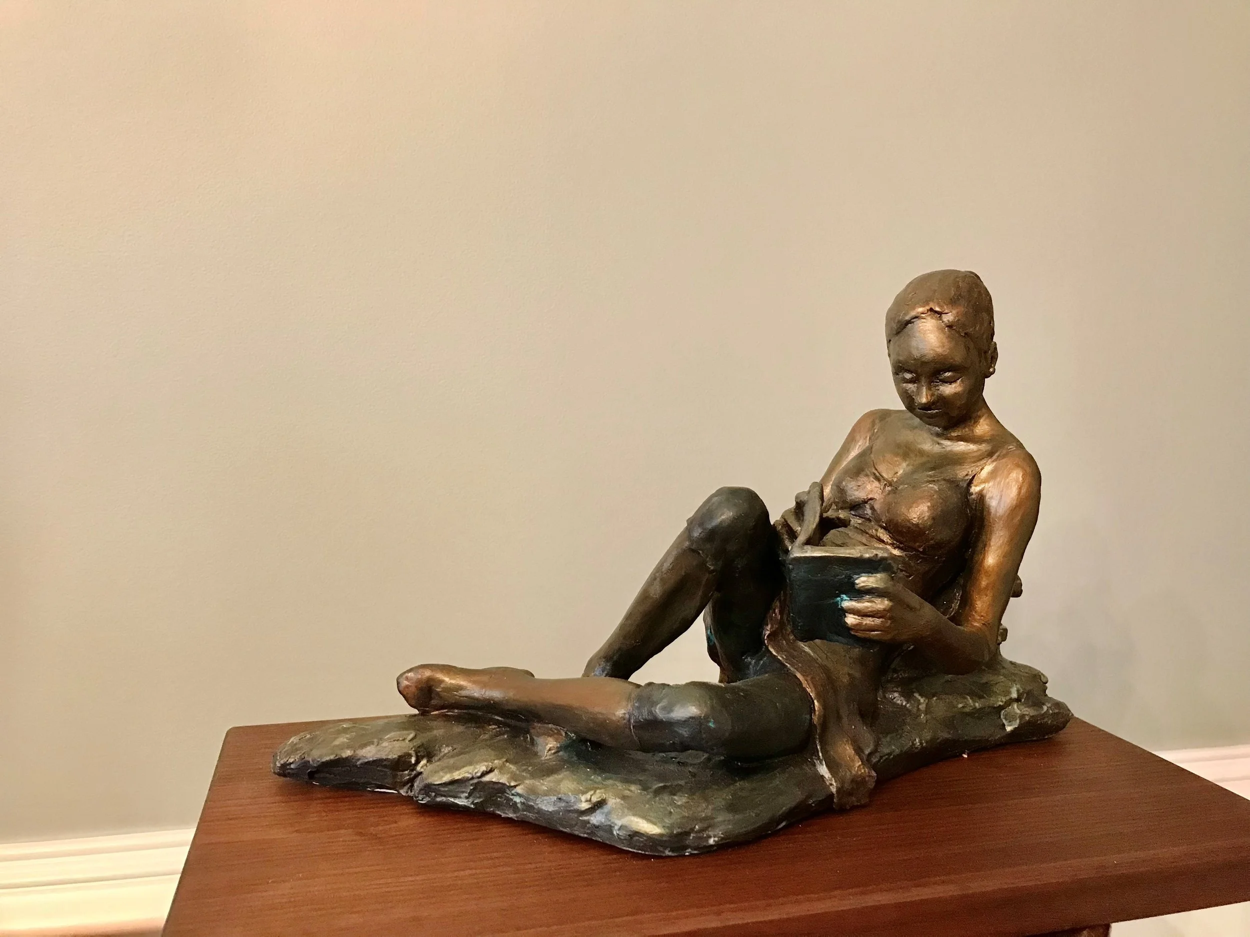 Bronze sculpture of a seated woman with crossed arms, sitting on a piece of driftwood, displayed on a wooden table against a plain wall.