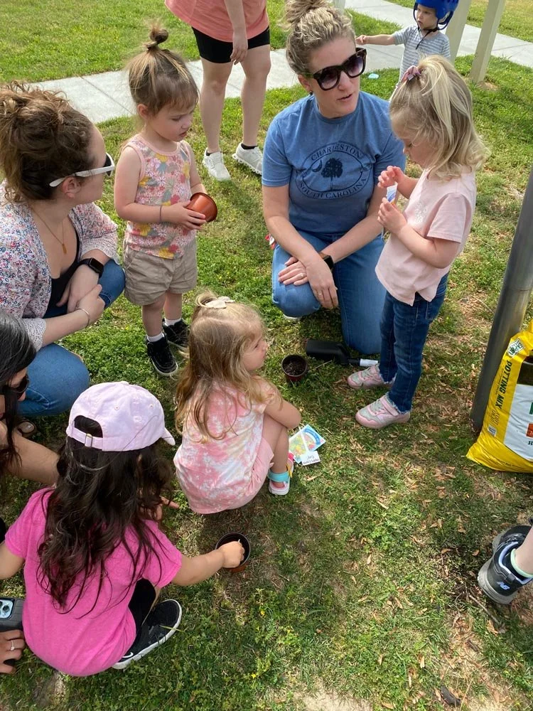 A group of children and women gathered outdoors on the grass, with some holding small containers, engaging in storytelling or playing.