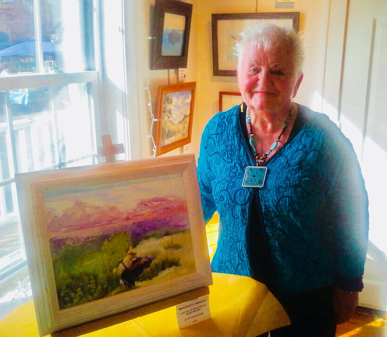 An elderly woman with short white hair wearing a blue jacket and colorful necklace standing in front of artwork in an art gallery or exhibition space.