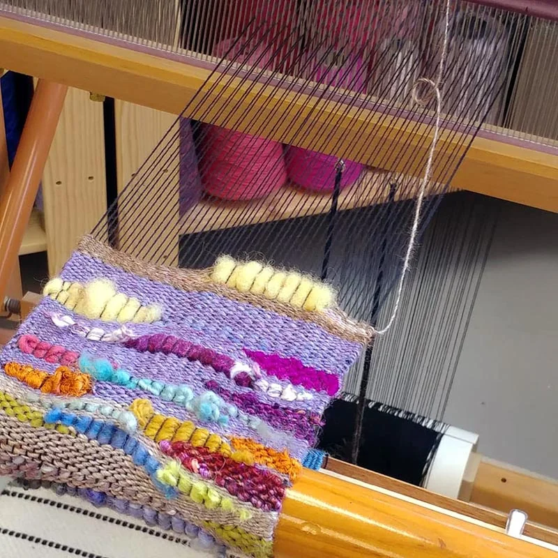 A loom weaving a colorful textile with various shades of purple, yellow, pink, blue, and orange yarns.