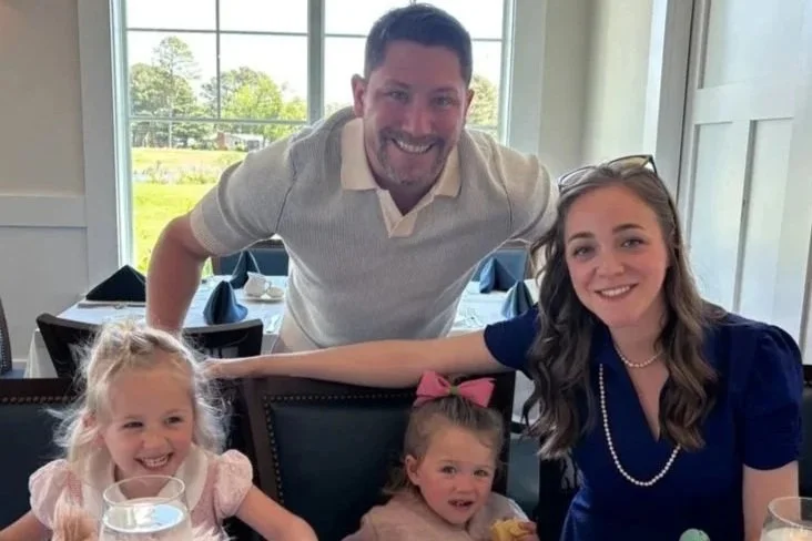 A man, woman, and two young girls sitting at a restaurant table, smiling and posing for a photo. The man is standing behind the girls, leaning over the back of the chair, while the women and girls sit at the table. There is a large window in the background with sunlight and greenery outside.