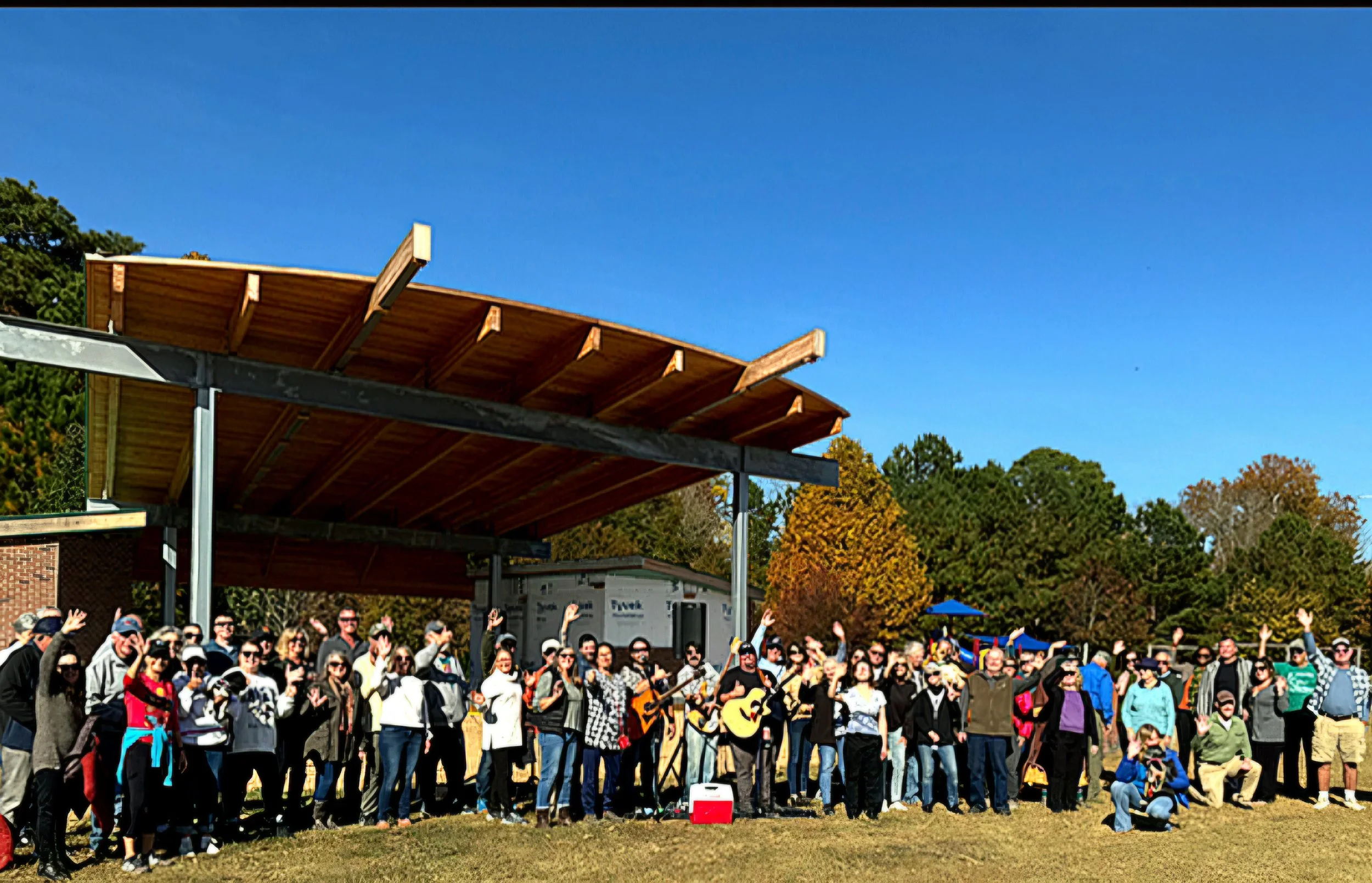A large group of people gathered outdoors in front of a stage with a wooden roof structure. The crowd is waving and wearing various colorful clothing, with some people taking photos. The sky is clear and blue, and there are trees with autumn foliage in the background.