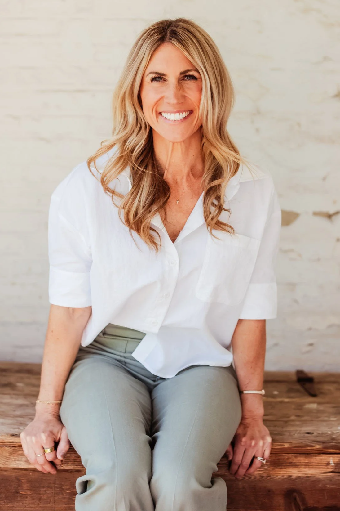 A smiling woman with blonde hair sitting on a wooden bench in front of a light-colored wall, wearing a white shirt and light-colored pants.