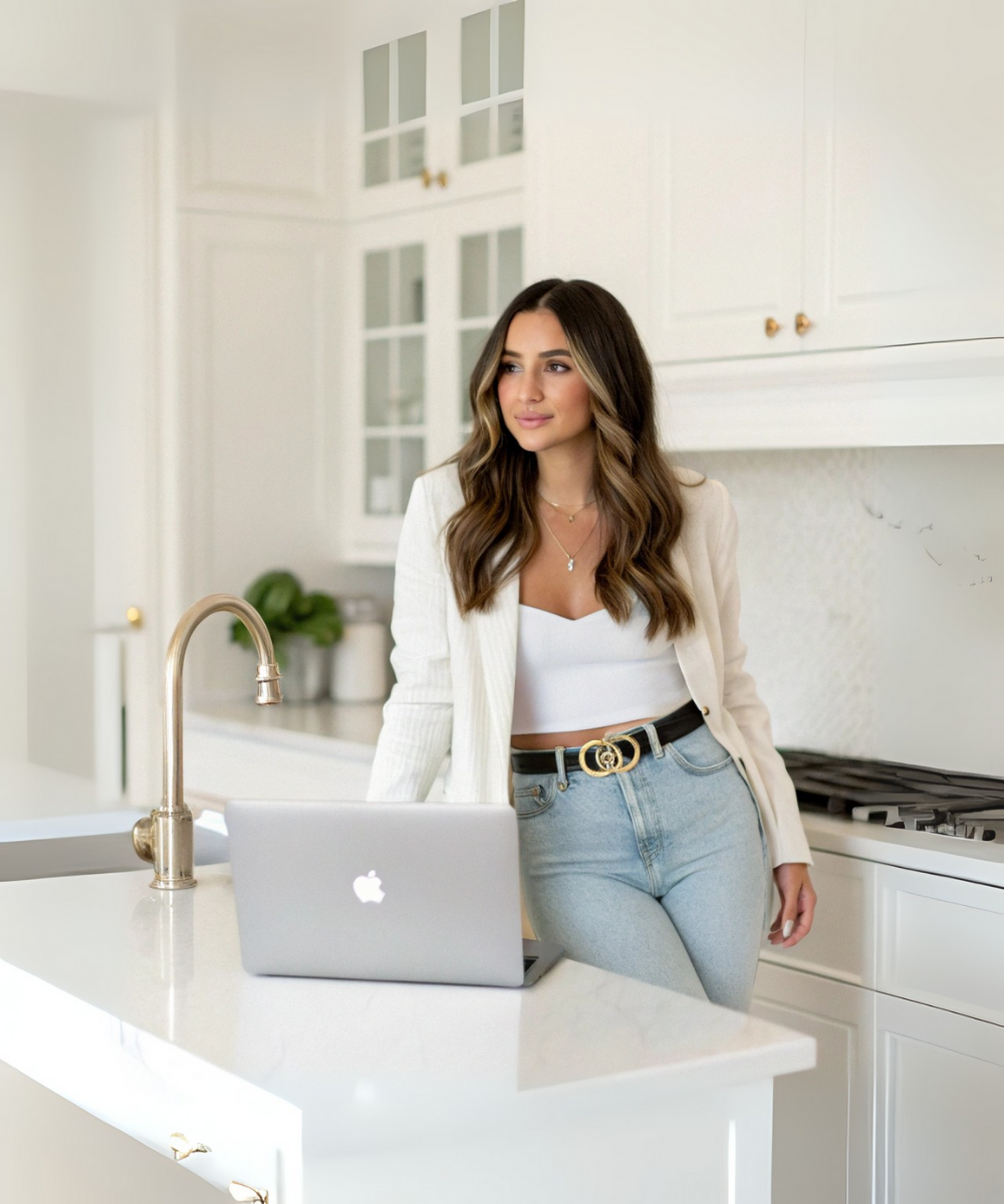 A woman with long brown hair, wearing a white cropped top, cream blazer, light blue jeans, and a blackbelt with gold details, stands in a modern white kitchen near a laptop on the counter.