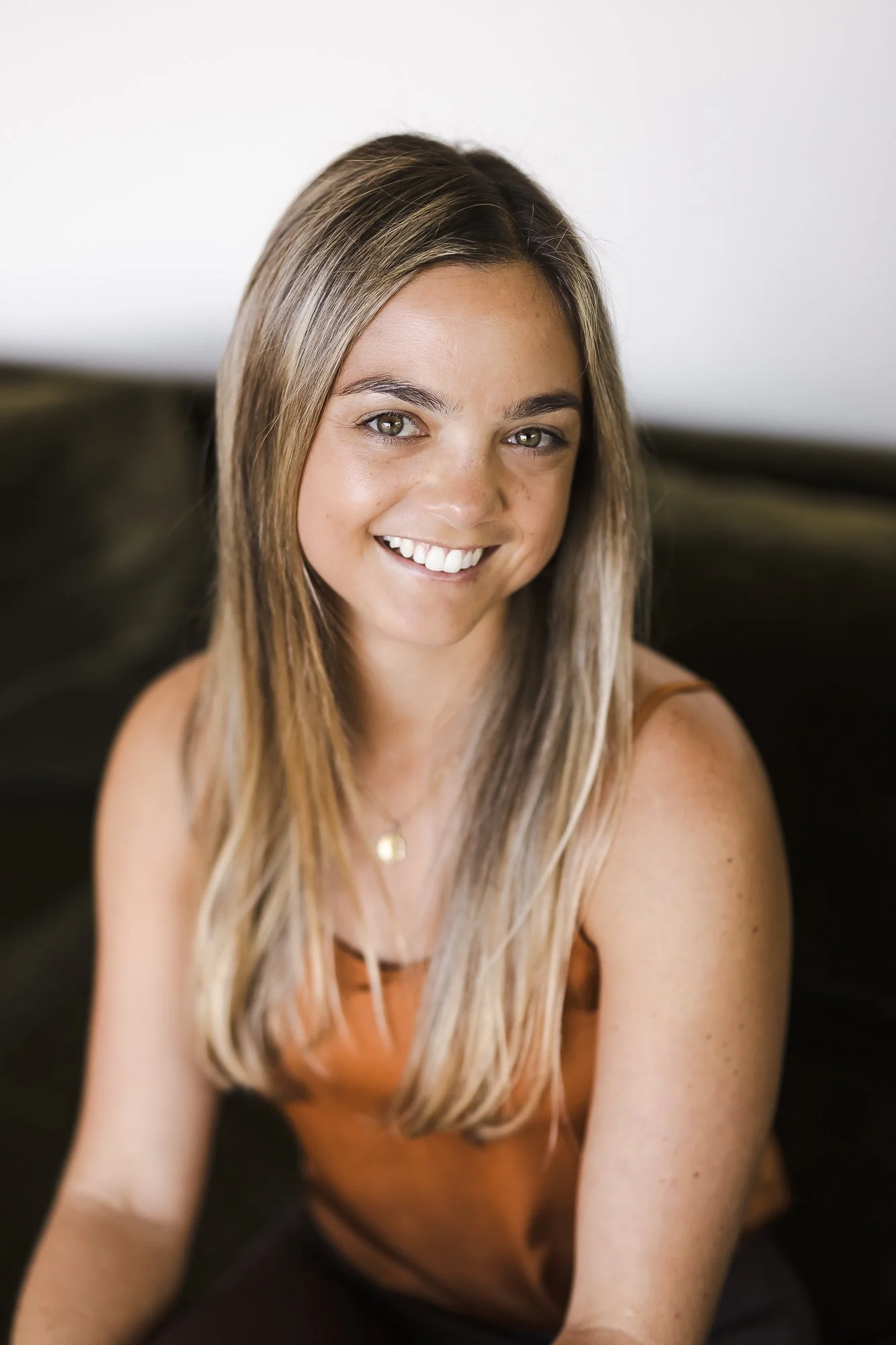 A young woman with long, blonde hair, smiling and looking at the camera, wearing an orange tank top and sitting on a dark couch with a plain, light-colored wall in the background.