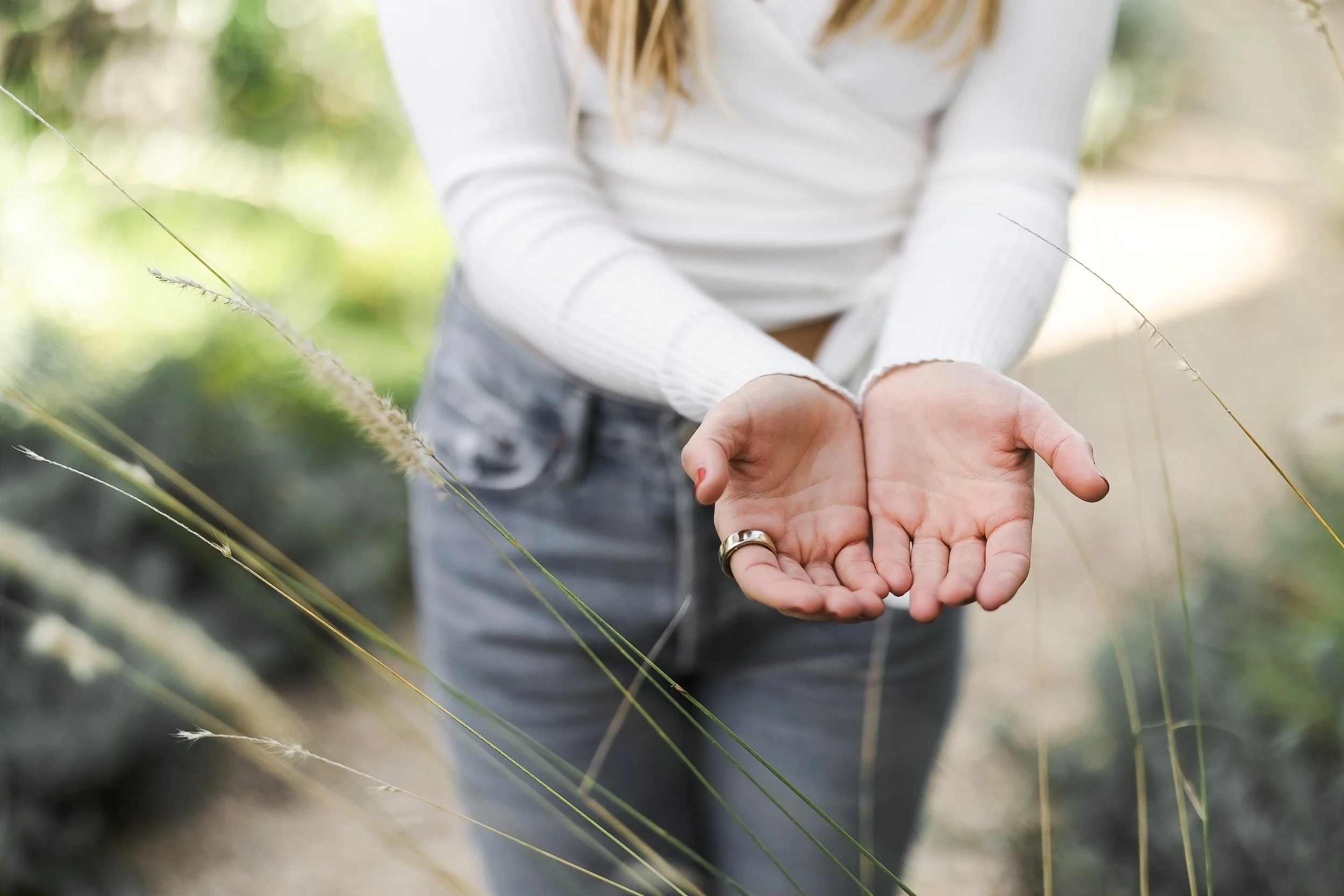 Person with long hair wearing a white sweater holding out open palms outdoors, surrounded by tall grasses and greenery.