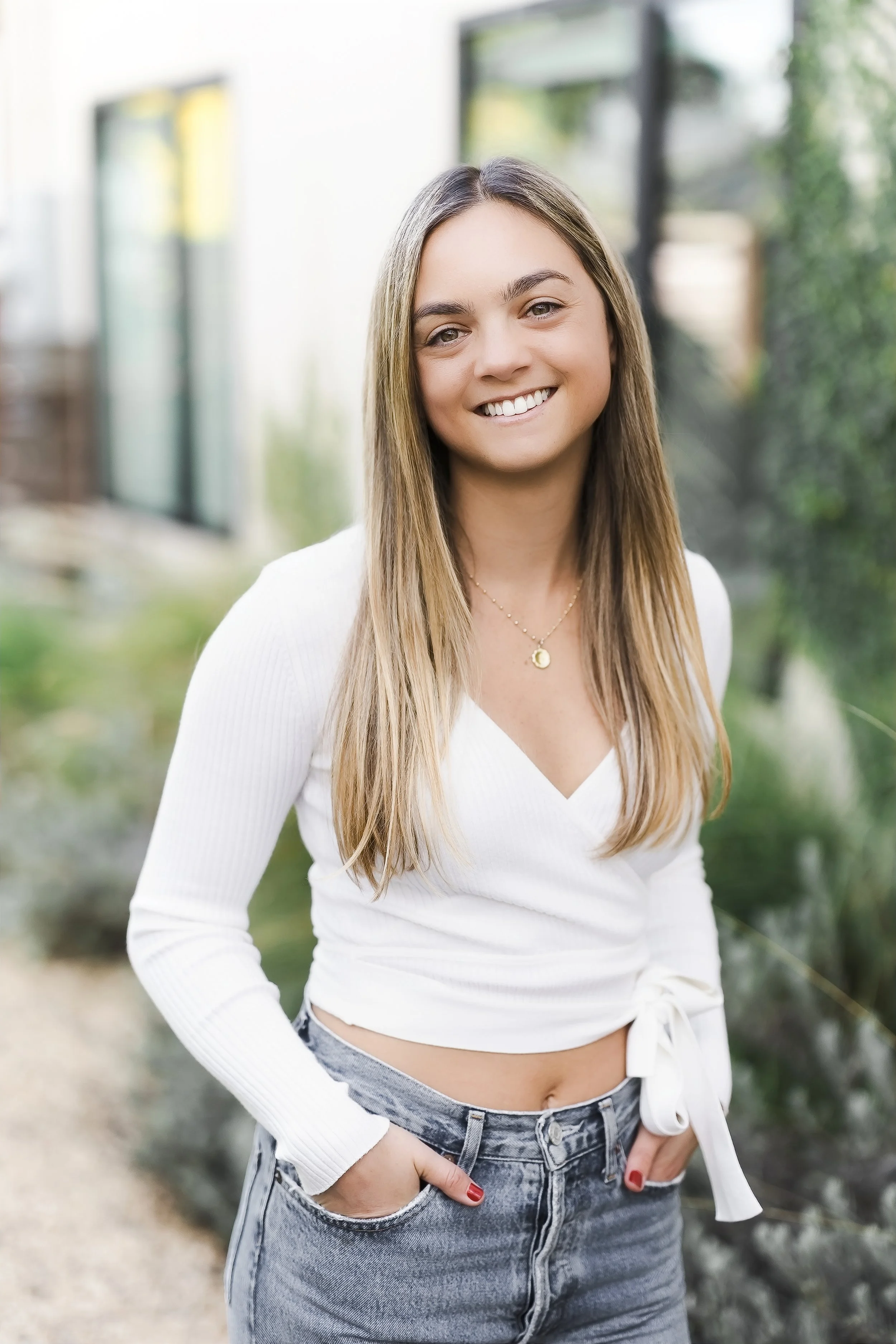 A young woman with long, light brown hair and a radiant smile, wearing a white cropped long sleeve top, high-waisted jeans, and a gold necklace, standing outdoors in front of greenery and windows.