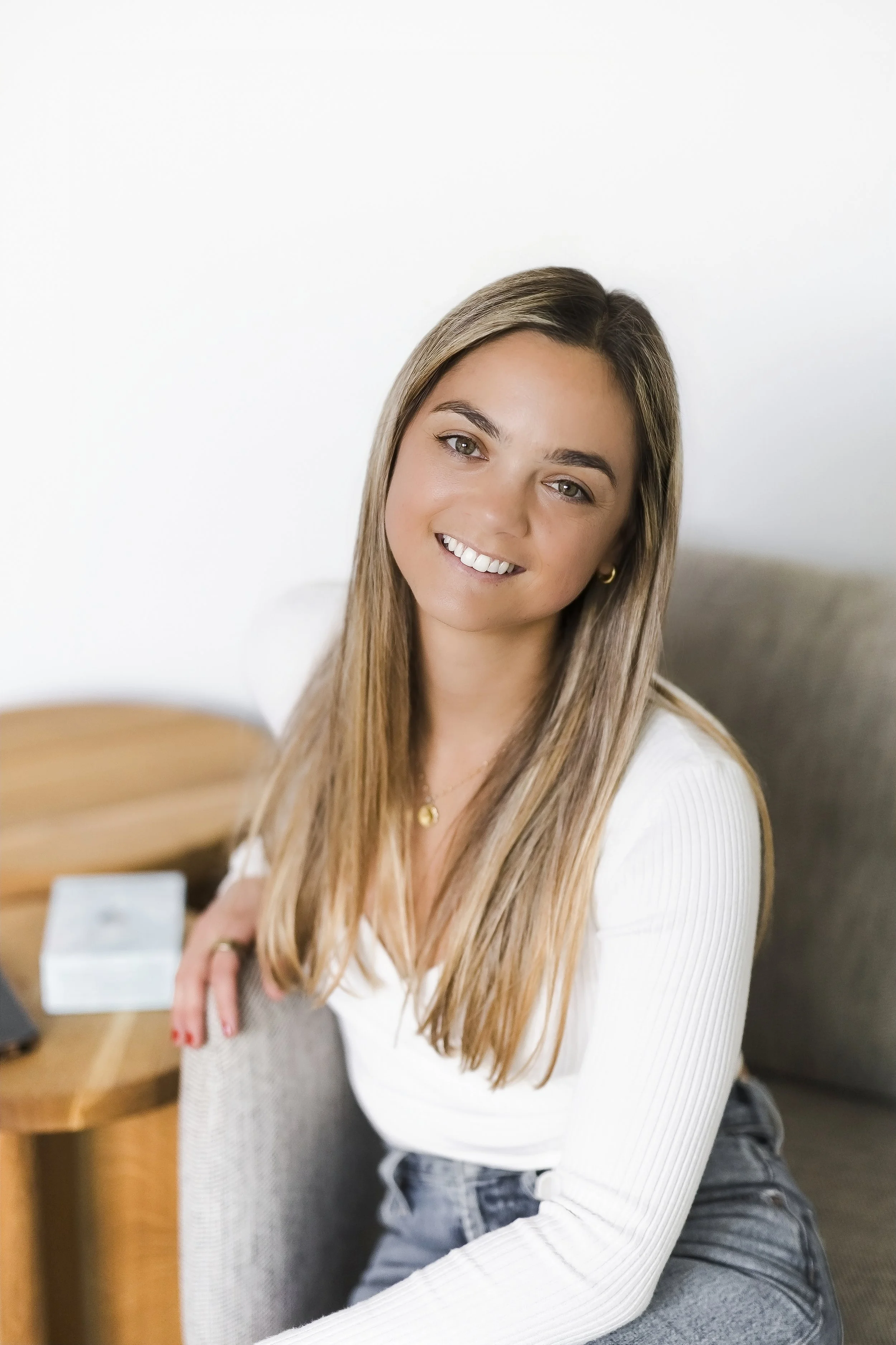 A young woman with long blonde hair, wearing a white long-sleeve top and gray jeans, smiling and leaning on a gray upholstered armchair in a bright room.