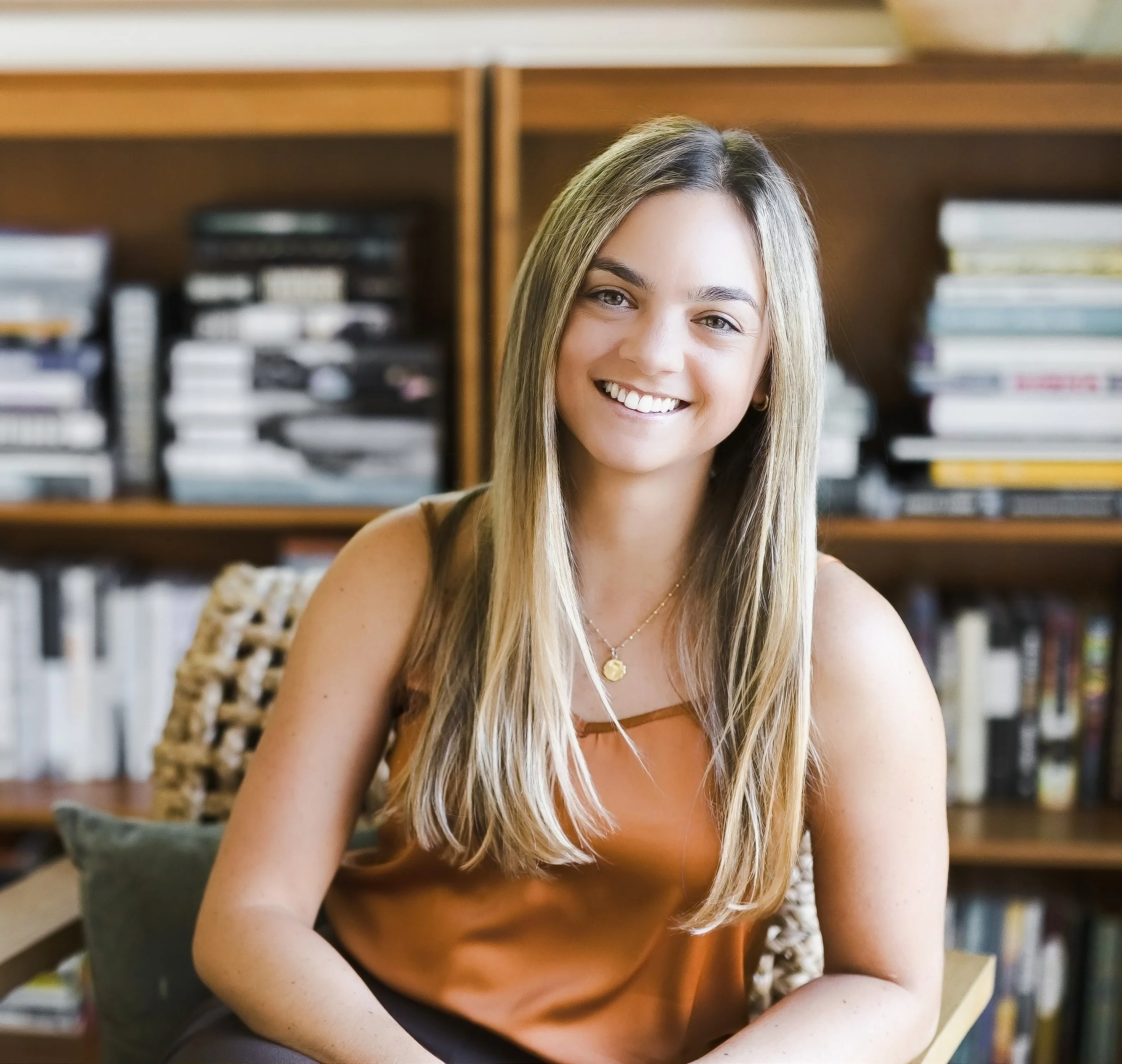 A woman with long blonde hair smiling and seated at a desk with bookshelves in the background.