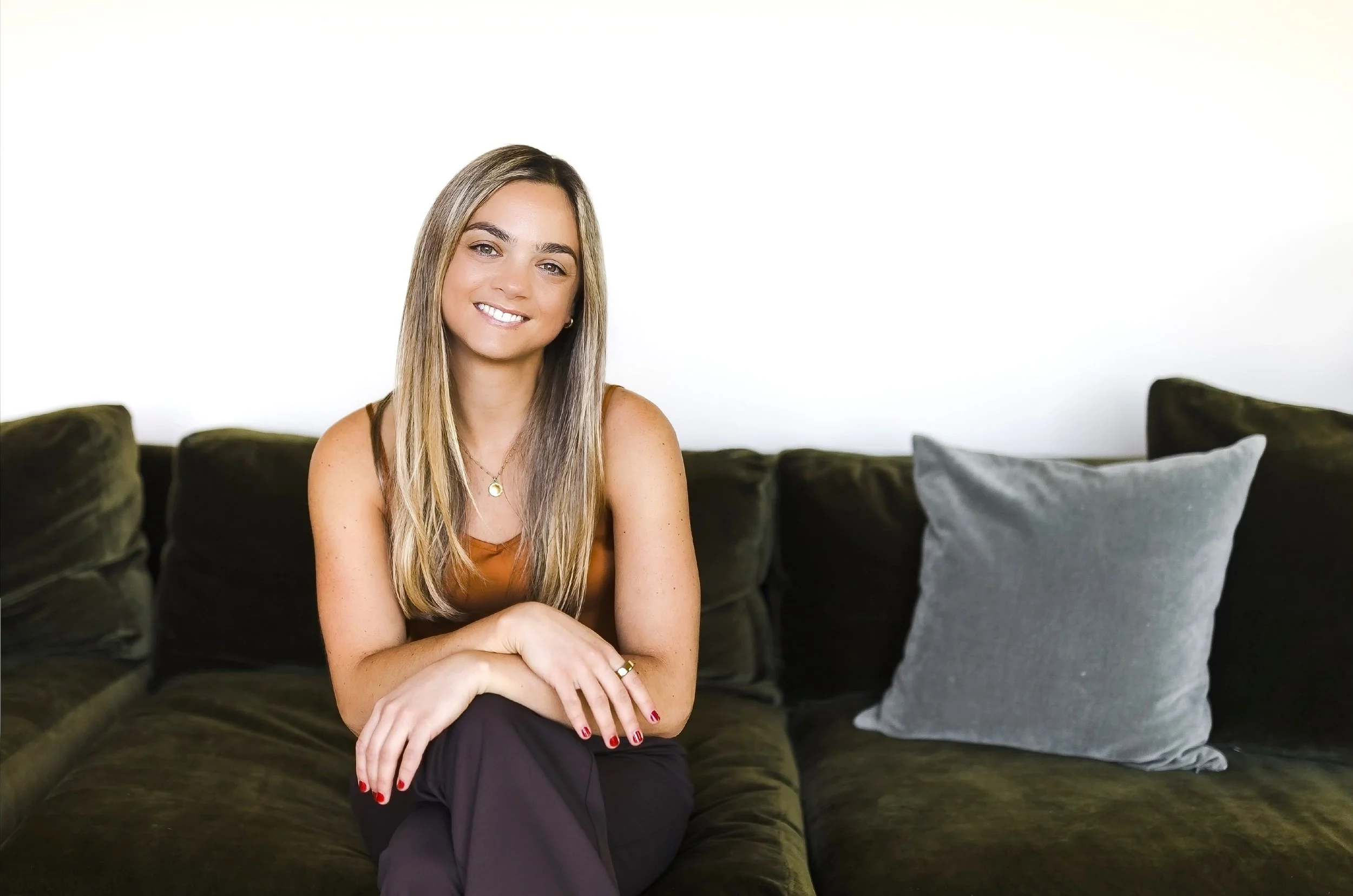 Stephanie Baubie, NP, smiling sitting on a dark green couch. She's wearing an Oura ring, a simple gold necklace, and her blond hair is long and straight.