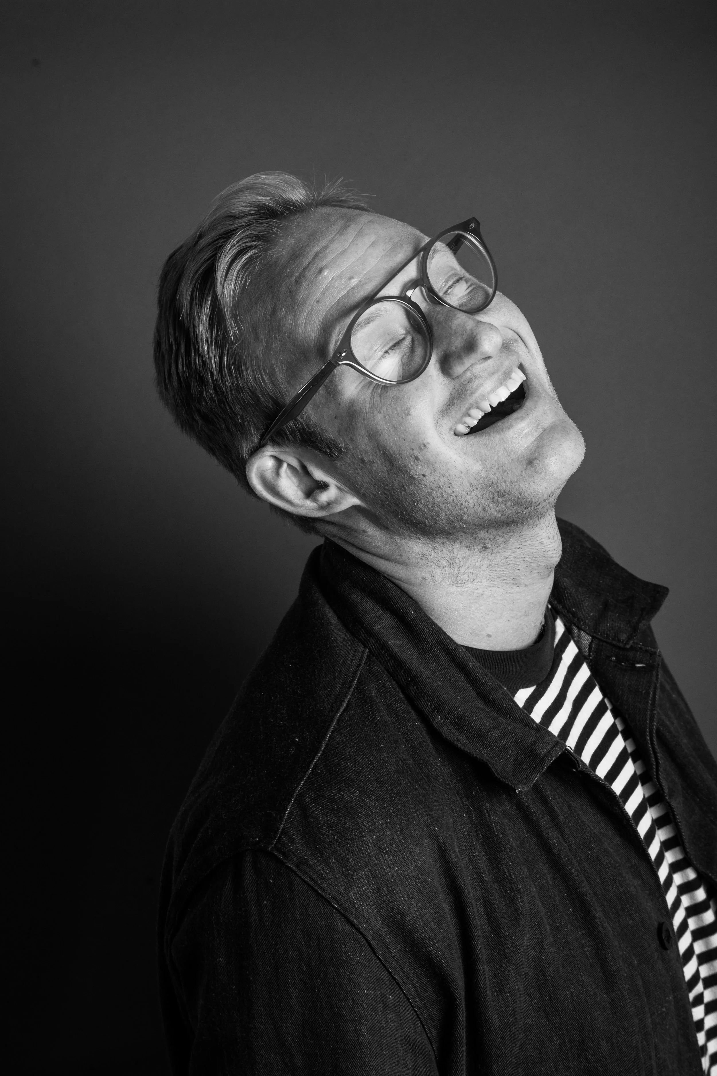 A black and white photo of a man laughing with eyes closed, wearing glasses, a striped shirt, and a dark jacket.