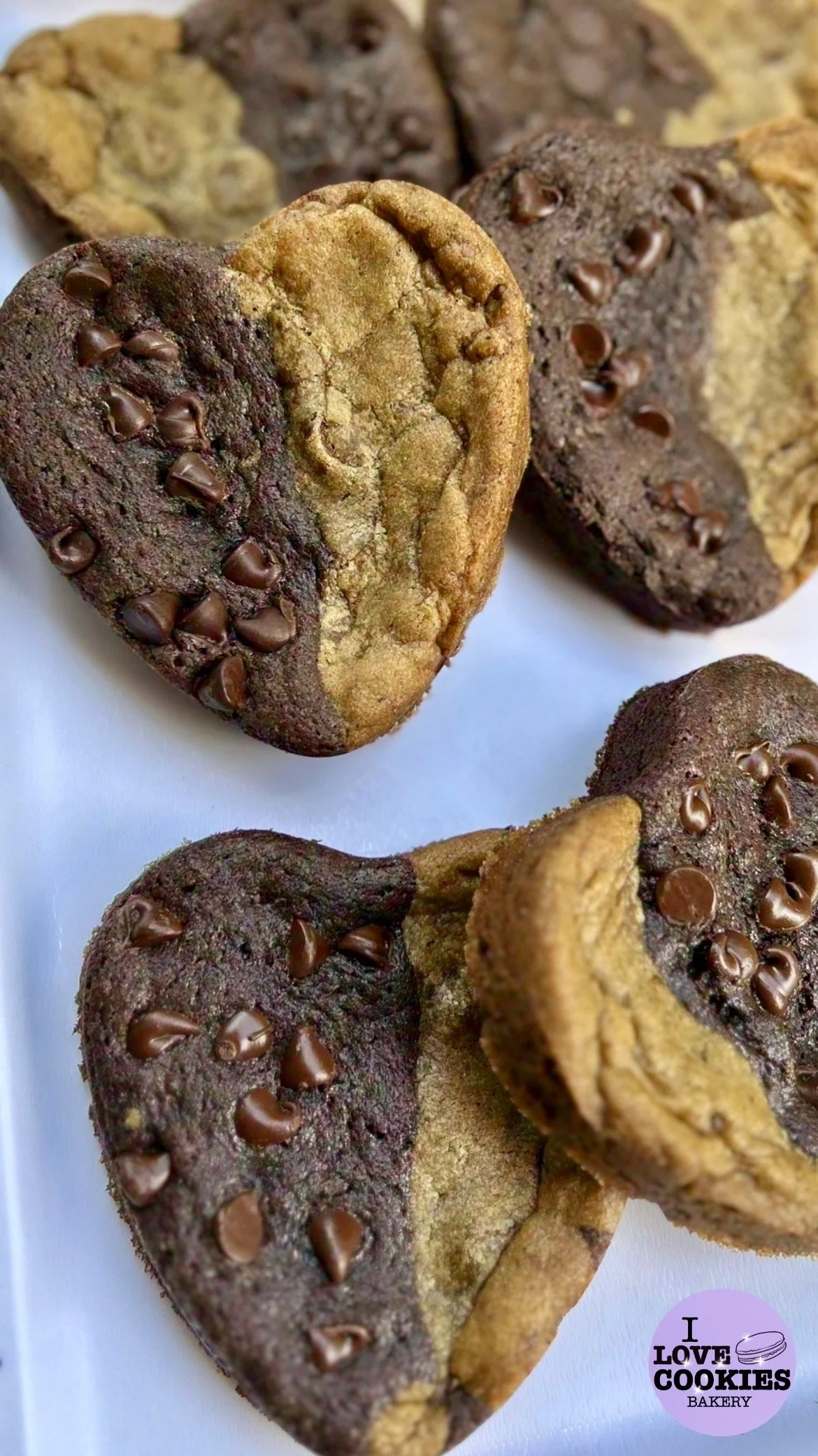 Heart-shaped cookies with a half chocolate and half vanilla appearance decorated with chocolate chips, placed on a white surface, with a purple bakery logo in the corner.