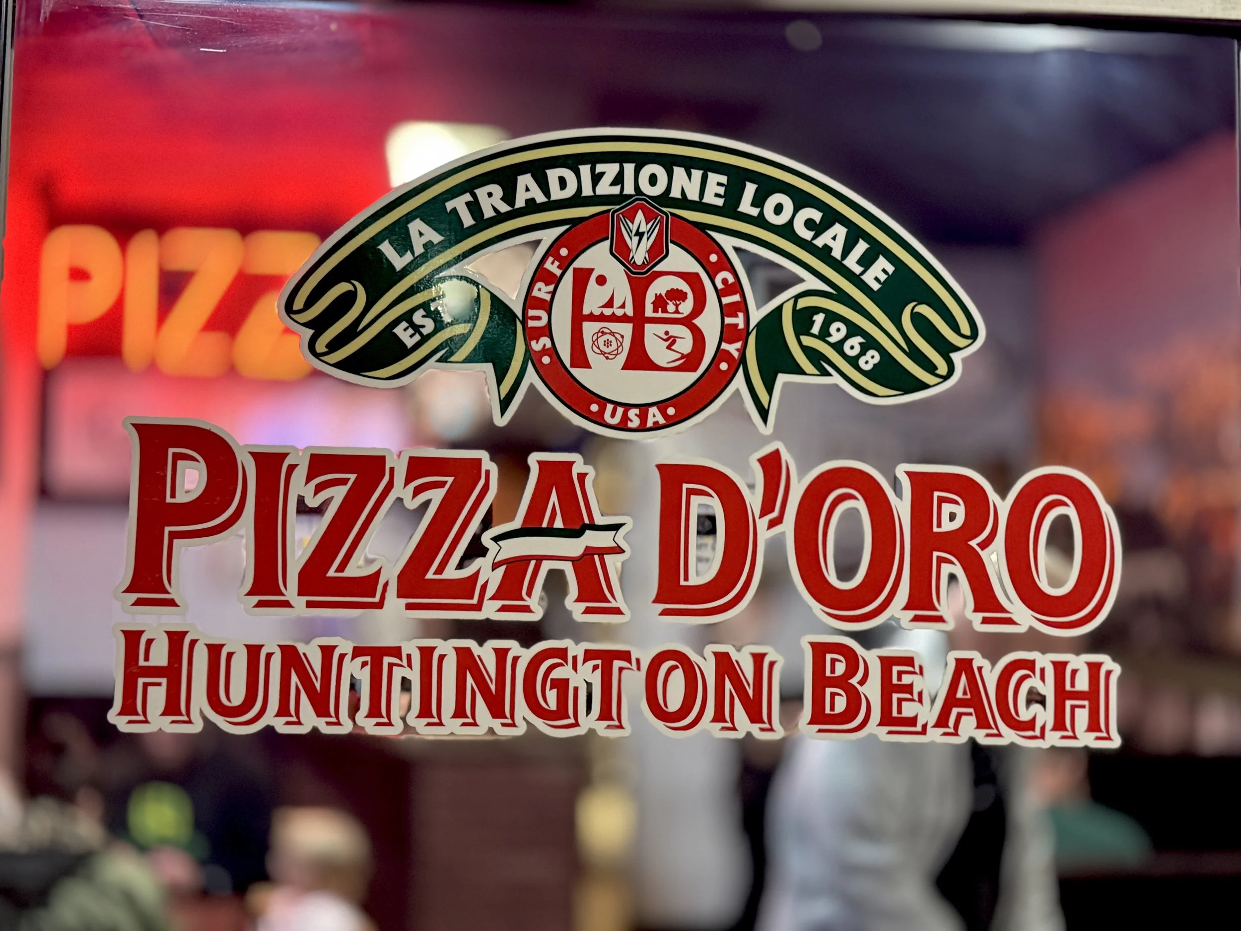 Colorful restaurant or storefront sign with the words 'Pizza D'Oro Huntington Beach' in red and white letters, and a circular emblem with various symbols and the text 'La Tradizione Locale,' 'Est. 1968,' and 'USA.'