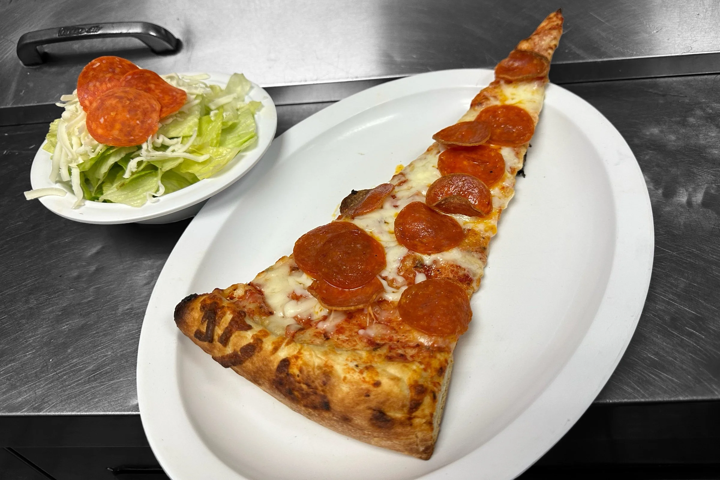 A slice of pepperoni pizza on a white plate and a small side salad with shredded cheese and lettuce, served on a metal surface.