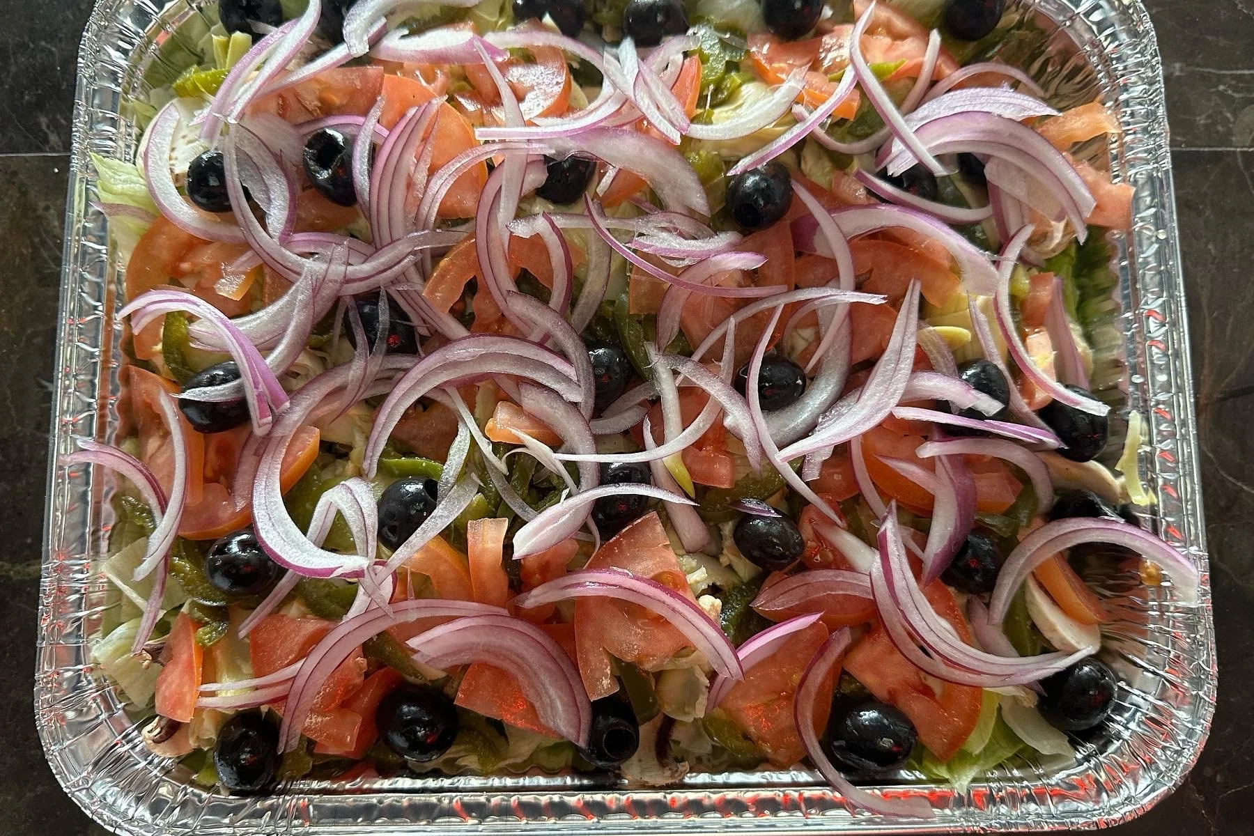 An aluminum tray filled with a salad consisting of sliced red onions, chopped tomatoes, green peppers, black olives, and shredded lettuce.