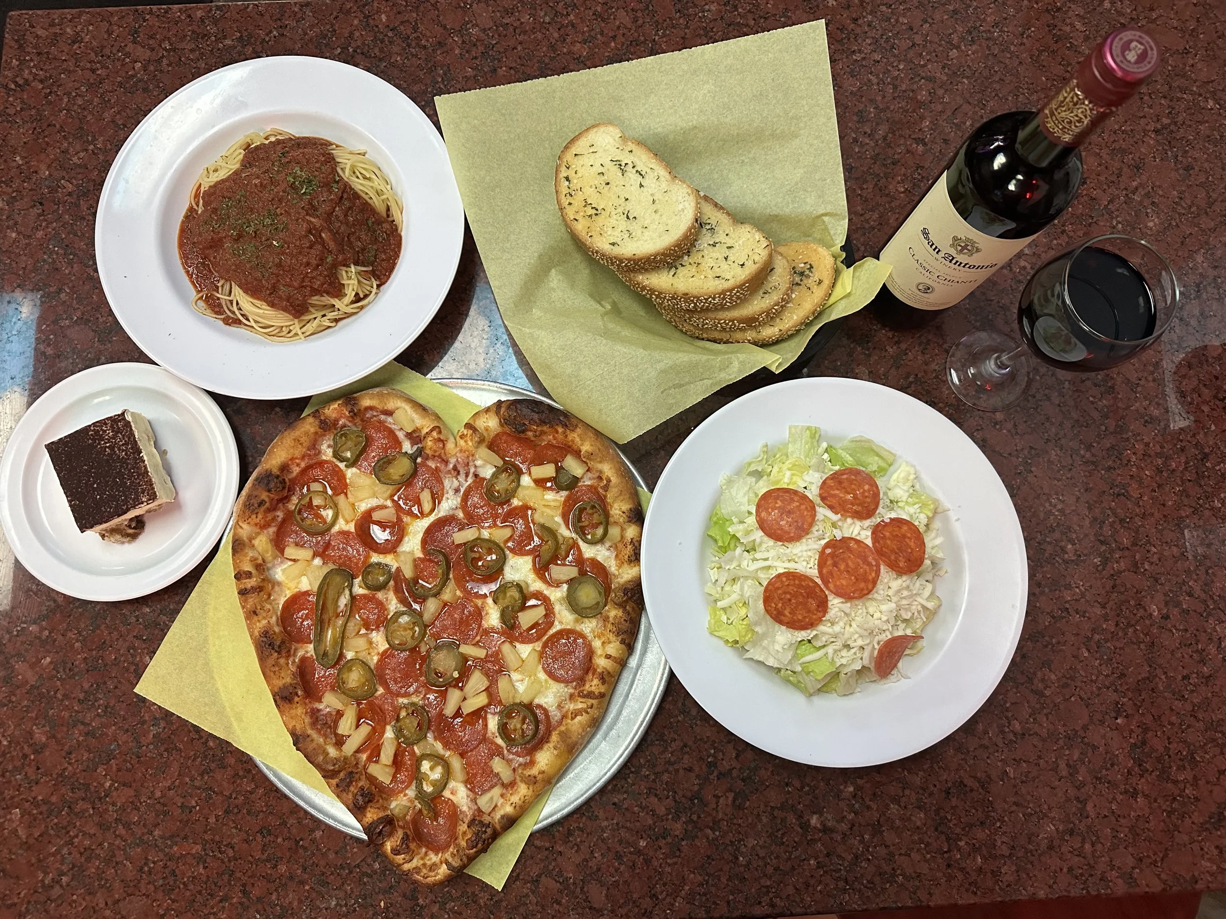 A meal including a large pepperoni and jalapeño pizza, garlic bread, a bowl of spaghetti, a slice of tiramisu, a bowl of salad, a bottle of red wine, and a glass of red wine.