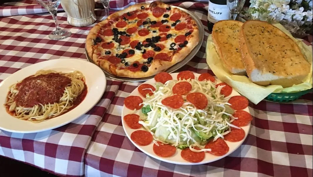 A table with a checkered red and white tablecloth set with a pepperoni and black olive pizza, a plate of spaghetti with marinara sauce, a salad with lettuce and pepperoni slices, two loaves of garlic bread, and two glasses of water.