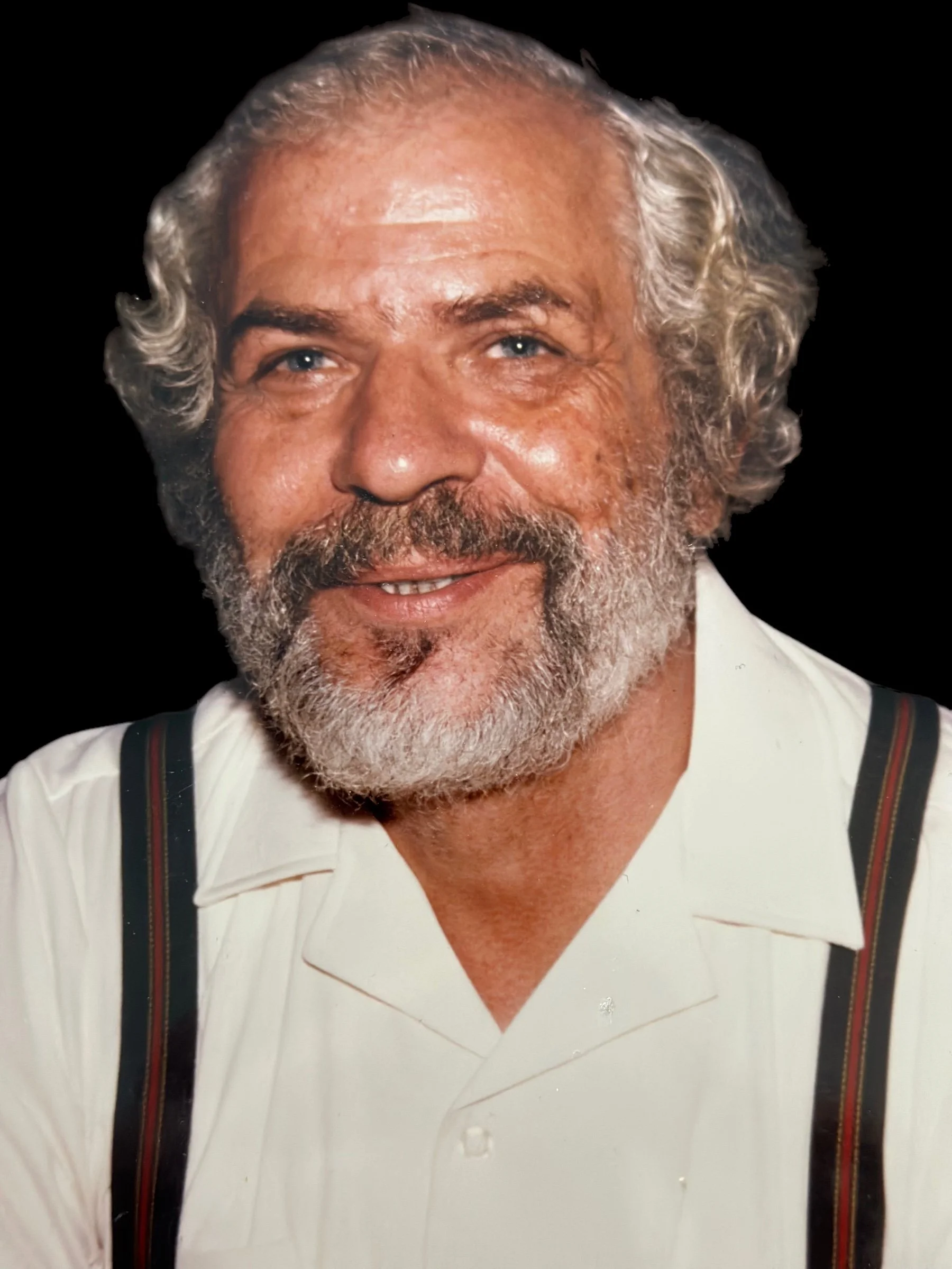 Close-up of Nick, our founder smiling with gray hair and beard wearing a white shirt and suspenders, against a black background.