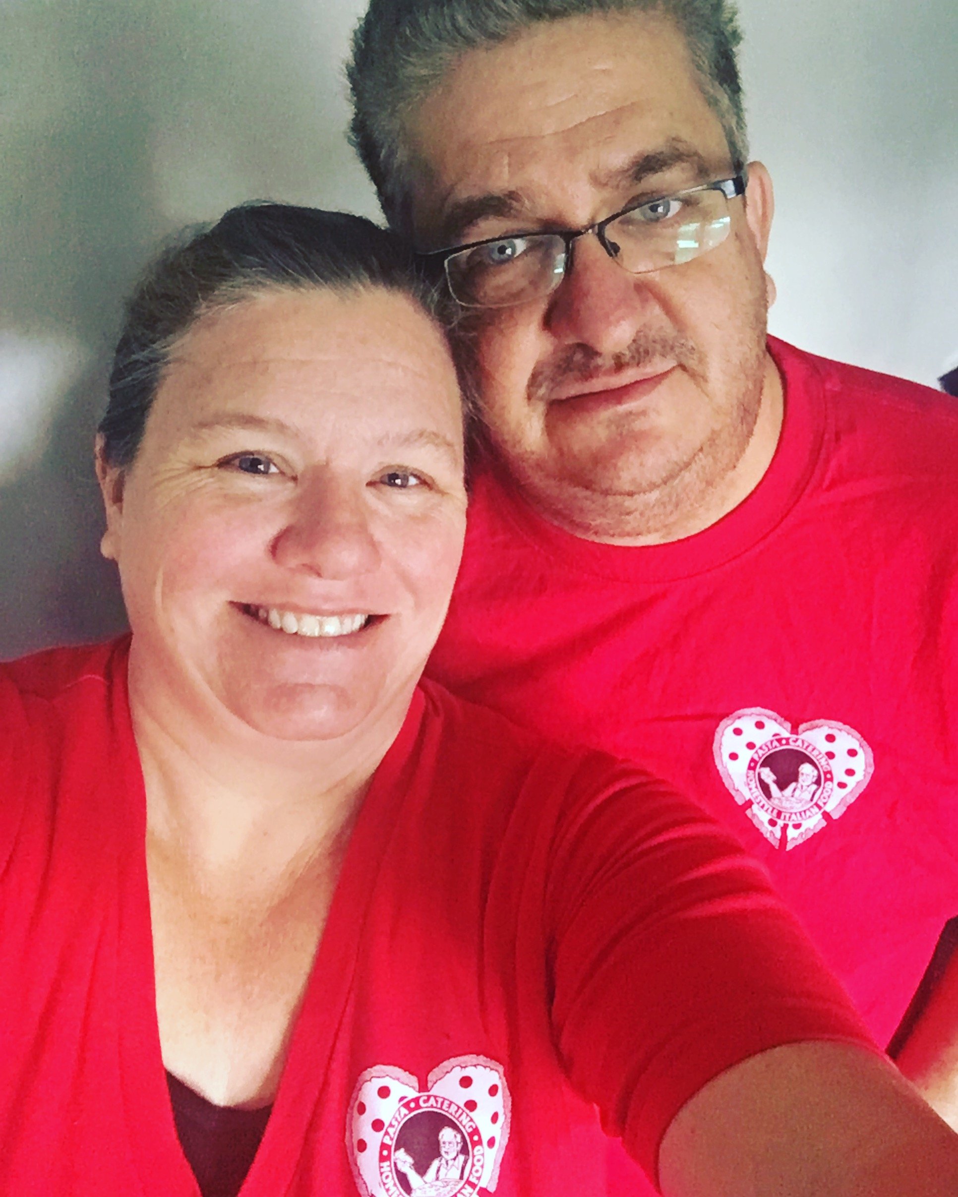 Close-up of Joe and Cindy wearing red shirts with a heart-shaped Pizza D'oro logo, standing indoors against a plain wall.