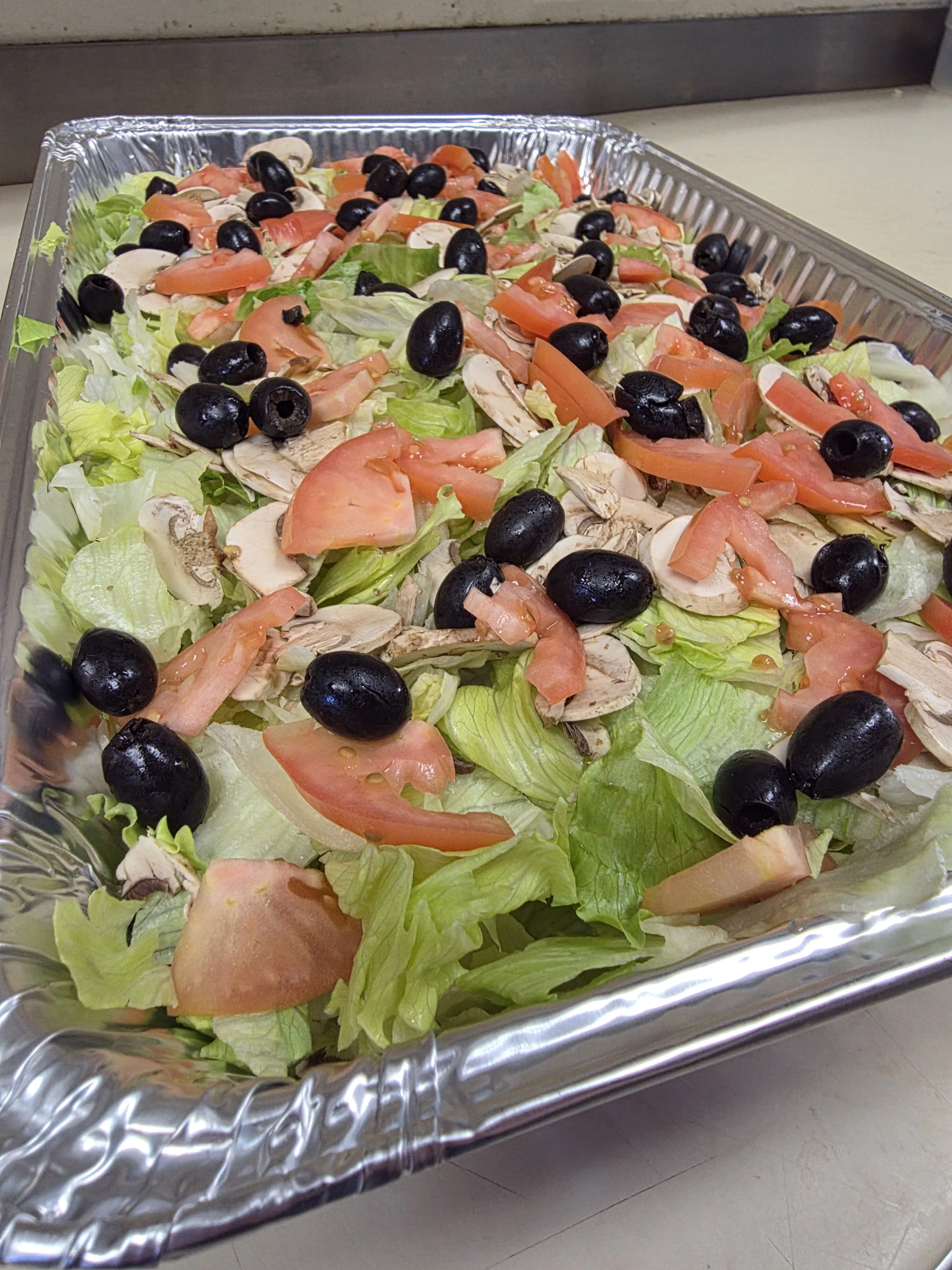 A tray of fresh garden salad with chopped lettuce, tomato slices, black olives, and sliced mushrooms in a metal container.