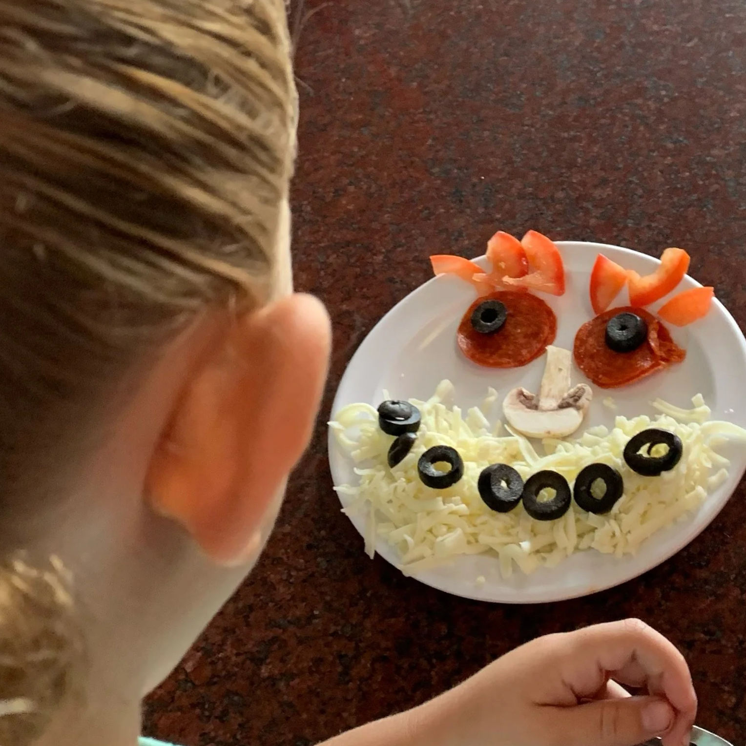 Child observing a plate with a face made of various foods, including sliced pepperoni and olives for eyes, shredded cheese for hair and face, sliced tomato pieces for hair, a mushroom and cheese for mouth.