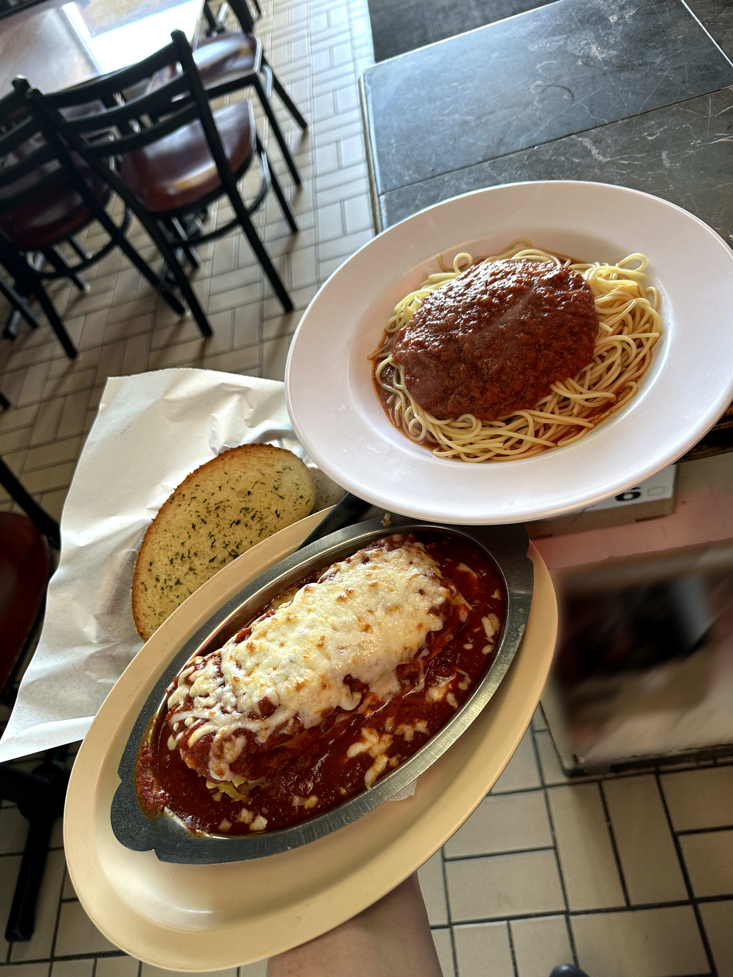 A plate of spaghetti with meat sauce, a baked Italian-style chicken Parmesan covered in melted cheese and marinara sauce, a garlic bread slice with herbs, all on a beige tray in a restaurant with black chairs and tiled floor.