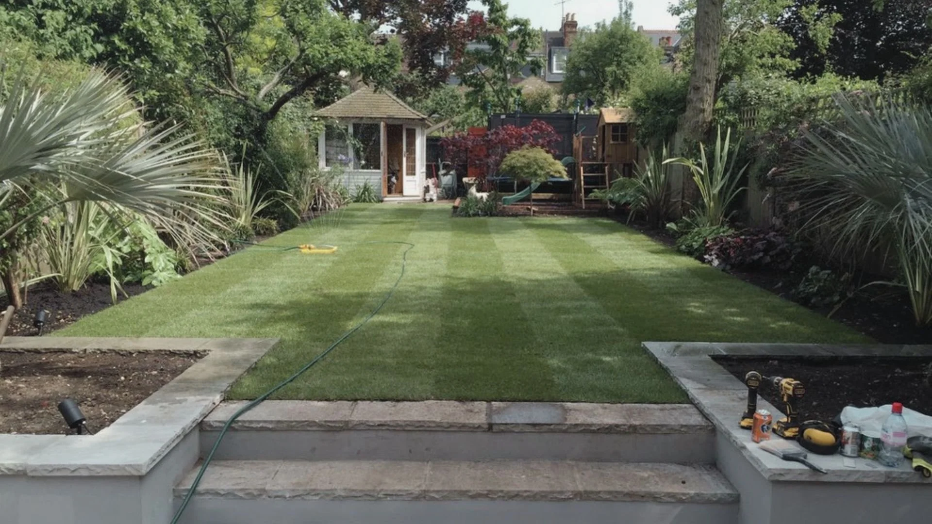 A backyard with a freshly laid green lawn, bordered by plants and trees, with a small garden shed and playhouse in the background. Tools and supplies are on the steps leading to the lawn, and a hose is laid out on the grass.