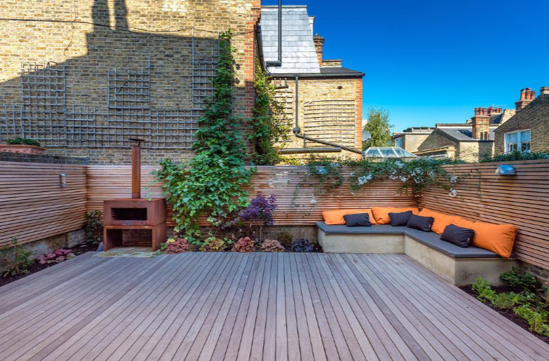Outdoor patio with wooden decking, built-in corner seating with orange and navy cushions, planter with climbing plants, and a red brick barbecue on a city rooftop or backyard, enclosed by wooden fencing and neighboring buildings visible in the background.