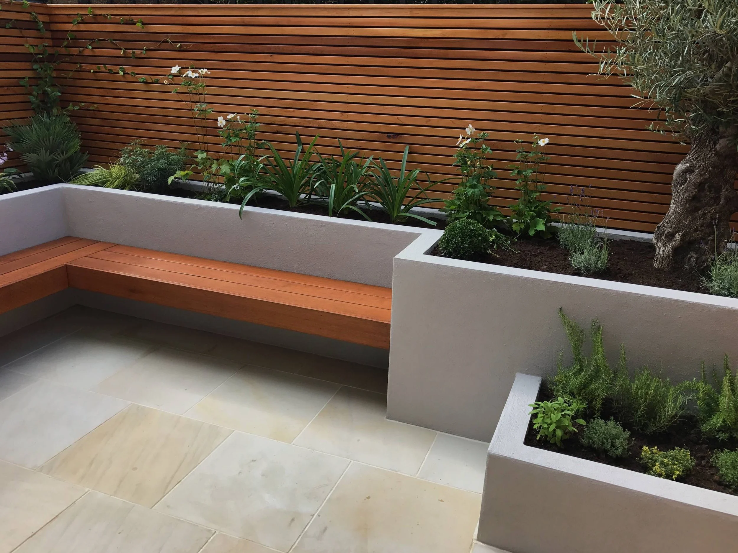 Modern outdoor patio with built-in wooden bench, concrete planters with various green plants, and a wooden slat fence in the background.