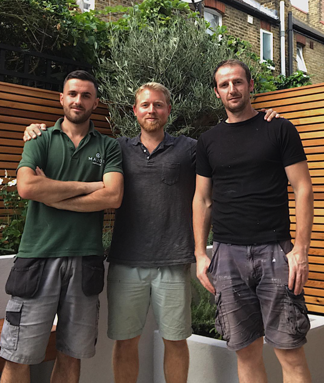 Three young men standing outdoors in front of a greenery wall and wooden fencing, posing together with relaxed expressions.