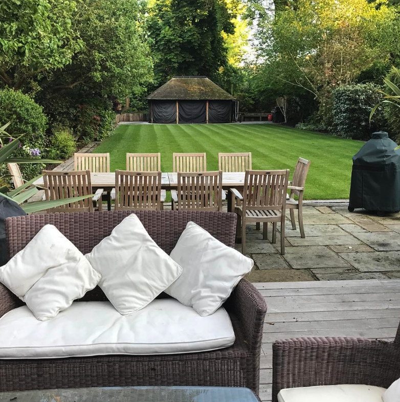 A backyard patio with a wicker sofa with white cushions and three white pillows, a dining table with eight wooden chairs, a well-maintained lawn, a small wooden shed, and lush green trees surrounding the area.