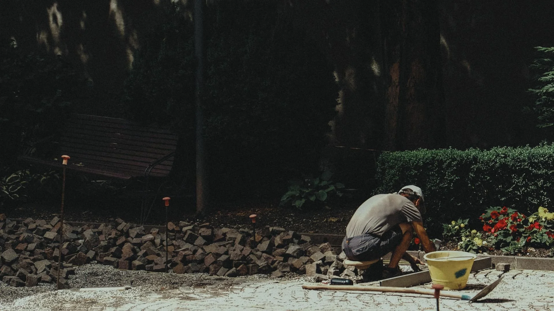 A person crouching and working on a sidewalk at night, surrounded by construction materials and tools, with plants and a park bench in the background.