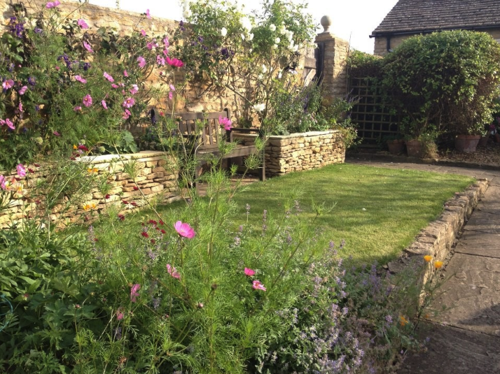 A cozy garden with a small lawn, flower beds, a stone wall with built-in seating, potted plants, and a trellis in the background, bathed in sunlight.