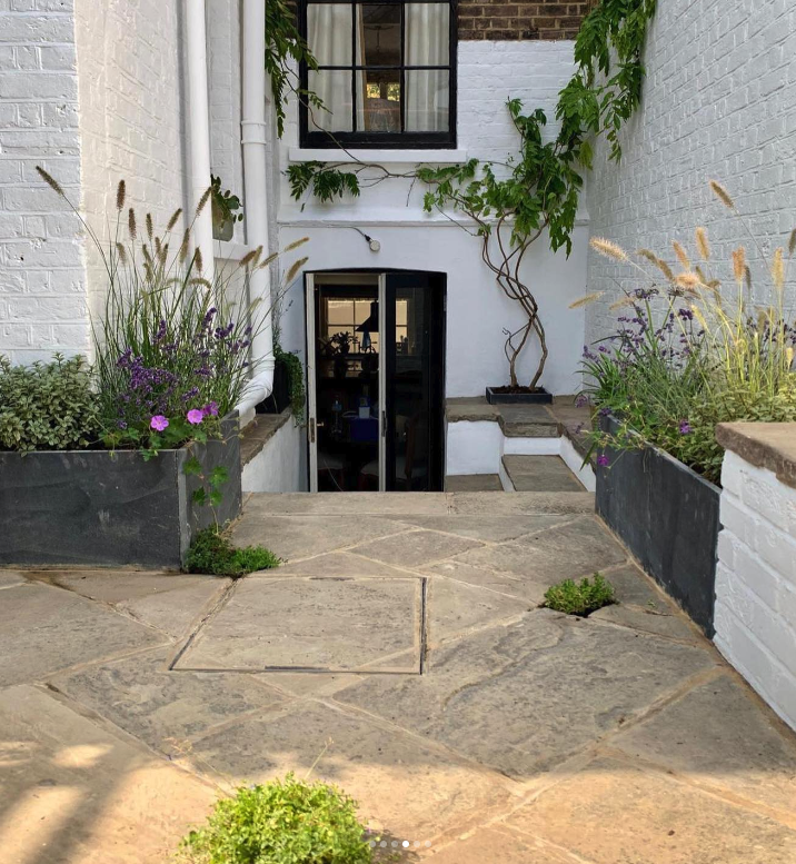 A small outdoor patio area with stone paving, two large planters with purple and green plants, and a white brick wall with a window and a door. There is also a twisting vine growing on the wall.