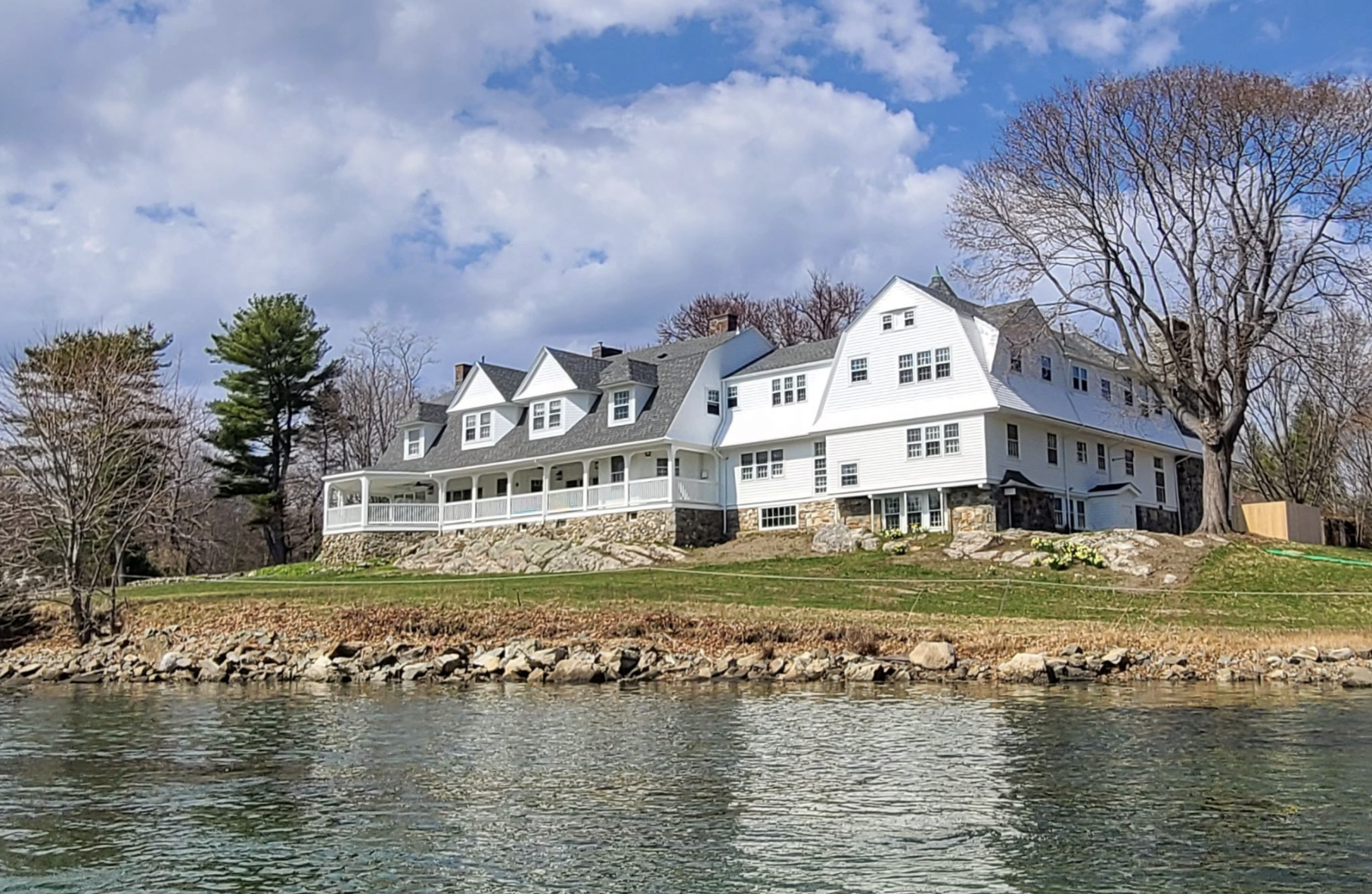 Large white house on a hill by a body of water, with trees and a partly cloudy sky in the background.