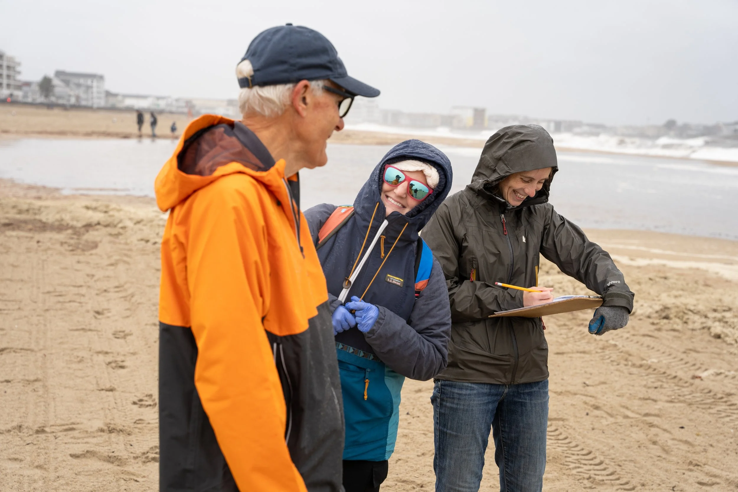 Three people in jackets and hats standing on a beach with buildings in the background, one woman is writing on a clipboard while the others look on, all smiling