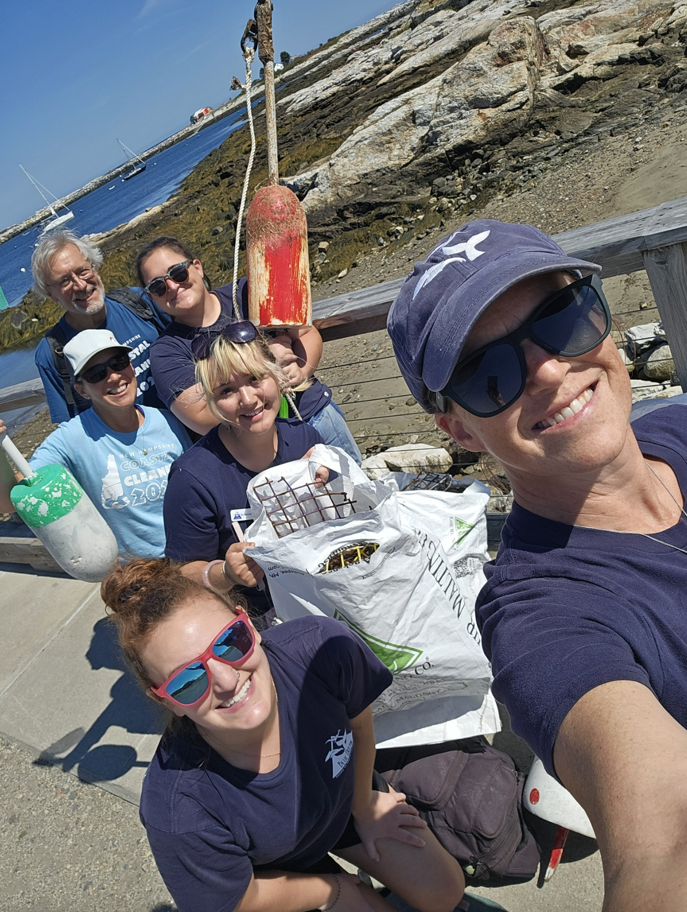A group of six people taking a selfie at the beach, with a rocky shoreline, a dock, boats, and a ship in the background on a sunny day.
