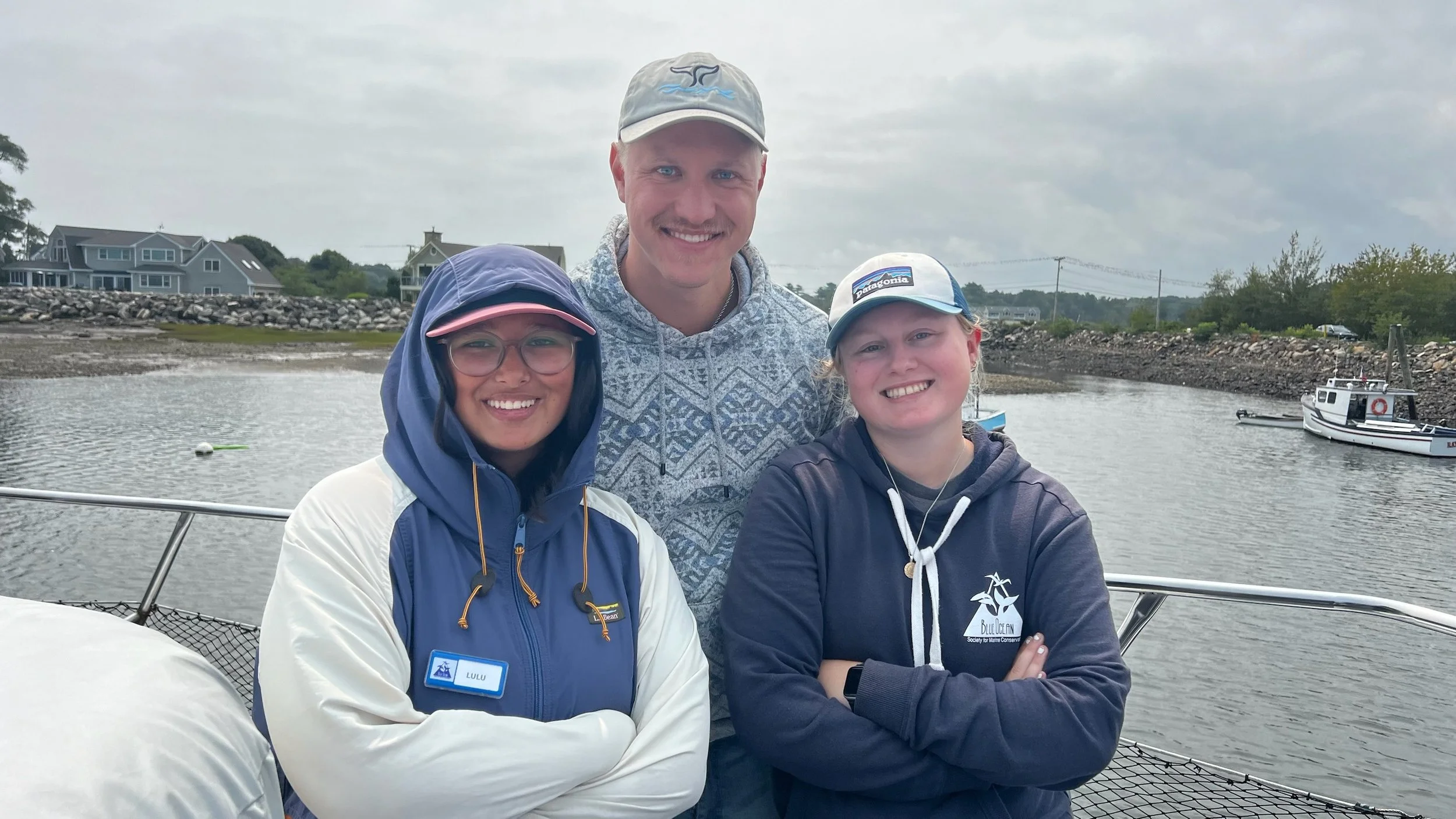 Three smiling people on a boat near a harbor, with houses and boats in the background, cloudy sky.