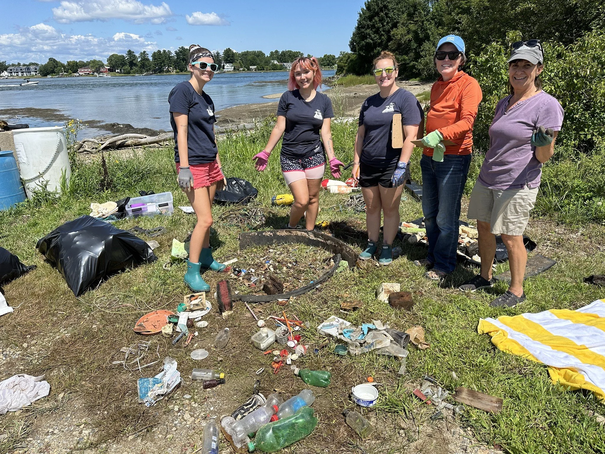 People standing on a grassy shoreline near a river, surrounded by trash including plastic bottles, cans, and bags, with trees and houses in the background.