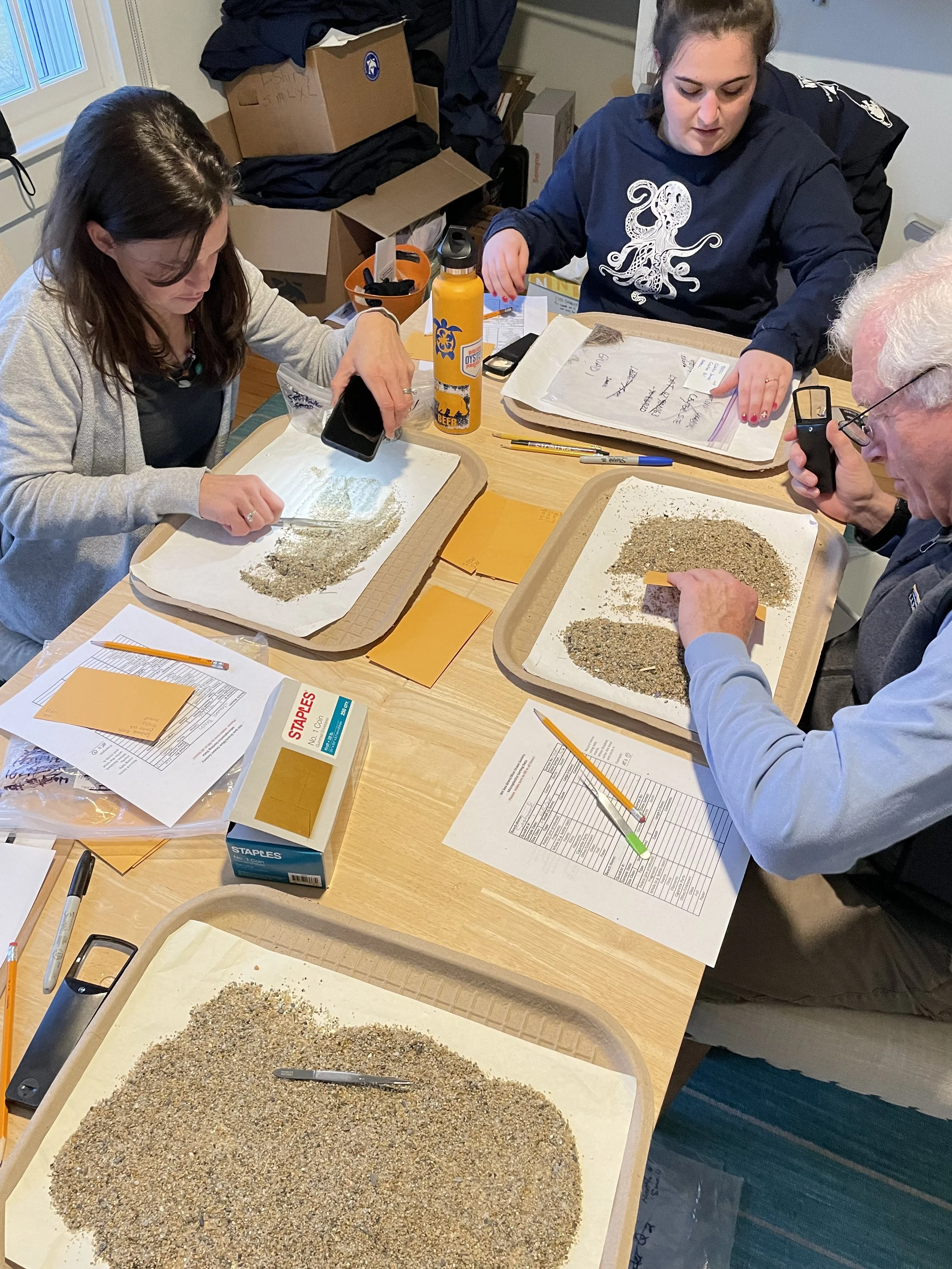 Four people sitting at a table examining and working with sand or soil on trays, with pens, papers, and office supplies on the table, surrounded by boxes and paperwork.
