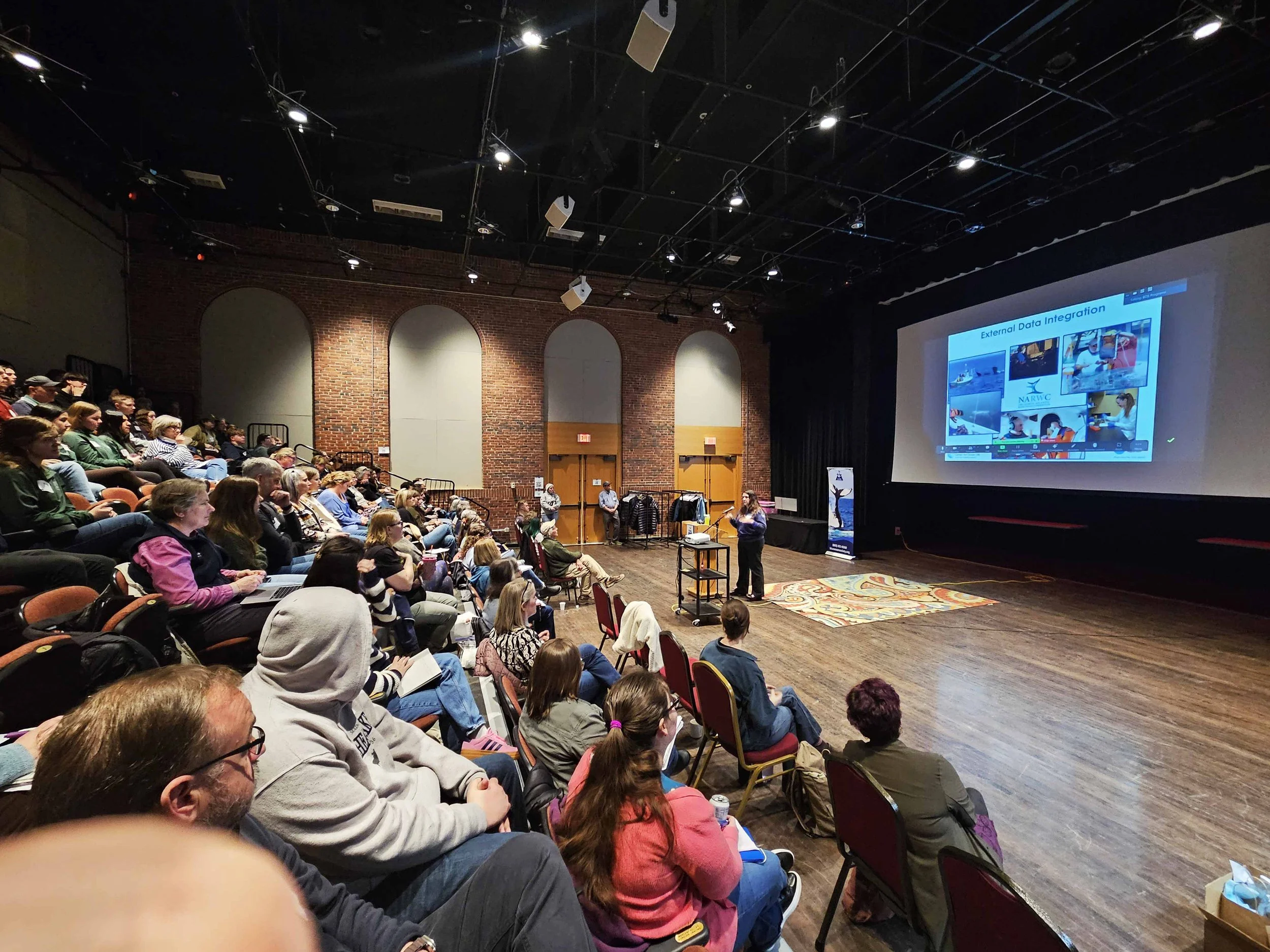 A large group of people sitting in an auditorium, attending a presentation or conference. The audience is looking towards a stage on the right side of the image, where a speaker is standing in front of a large screen displaying a slide titled 'External Data Integration' with various images and text. The room has a brick wall on the back with three arched windows, and the ceiling has black exposed beams and lighting equipment.