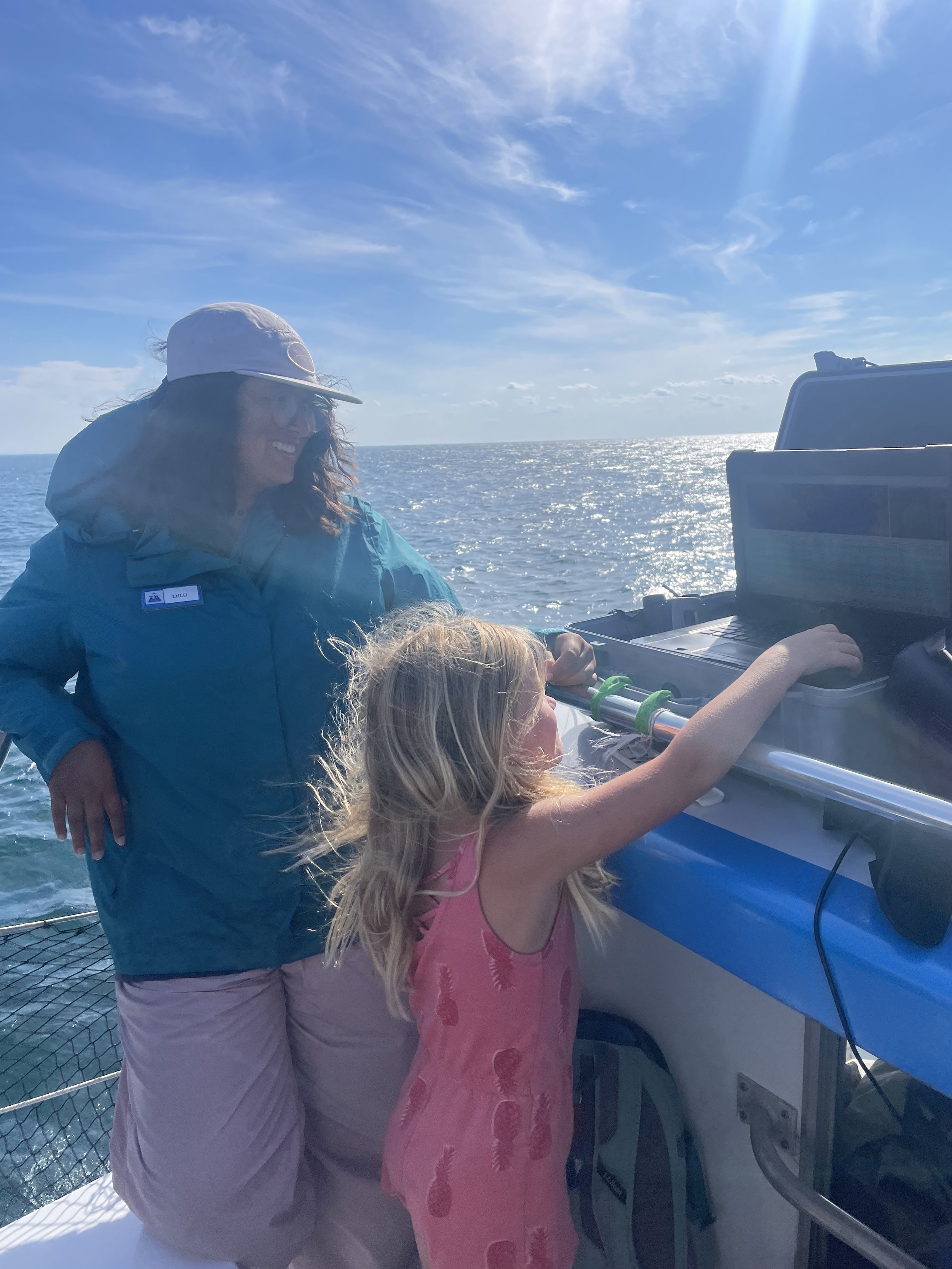 A woman with curly hair and wearing a white cap and glasses stands next to a young girl with blonde hair in a pink dress on a boat, both looking at equipment while sailing on the ocean with a bright blue sky and sunlight overhead.