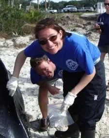 Two people, a woman and a boy, smiling and working together outdoors. The woman is wearing sunglasses and gloves, and they appear to be involved in a cleanup or maintenance activity near a rocky or sandy area.