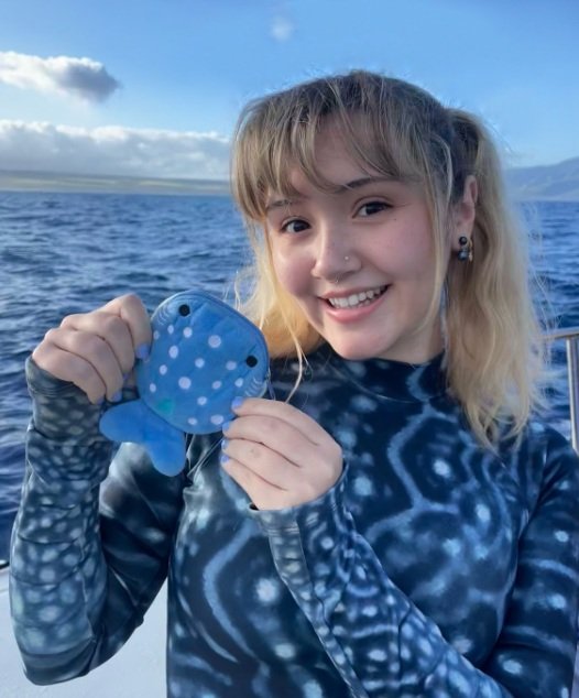 Young girl with blonde hair and earrings holding a blue fish-shaped object with white polka dots on a boat, with water and the sky in the background.