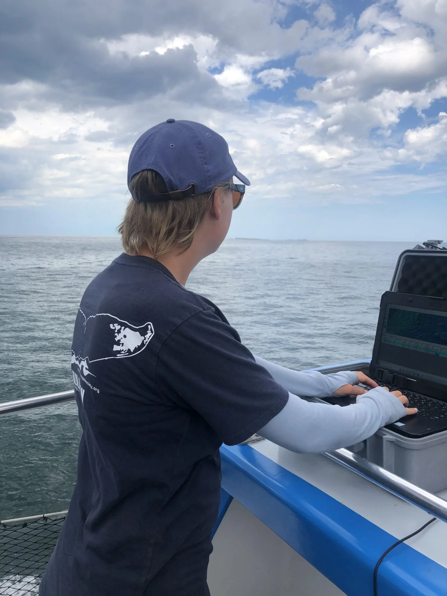 A person wearing a navy blue cap and sunglasses operating a radar or sonar device on a boat, with water and cloudy sky in the background.