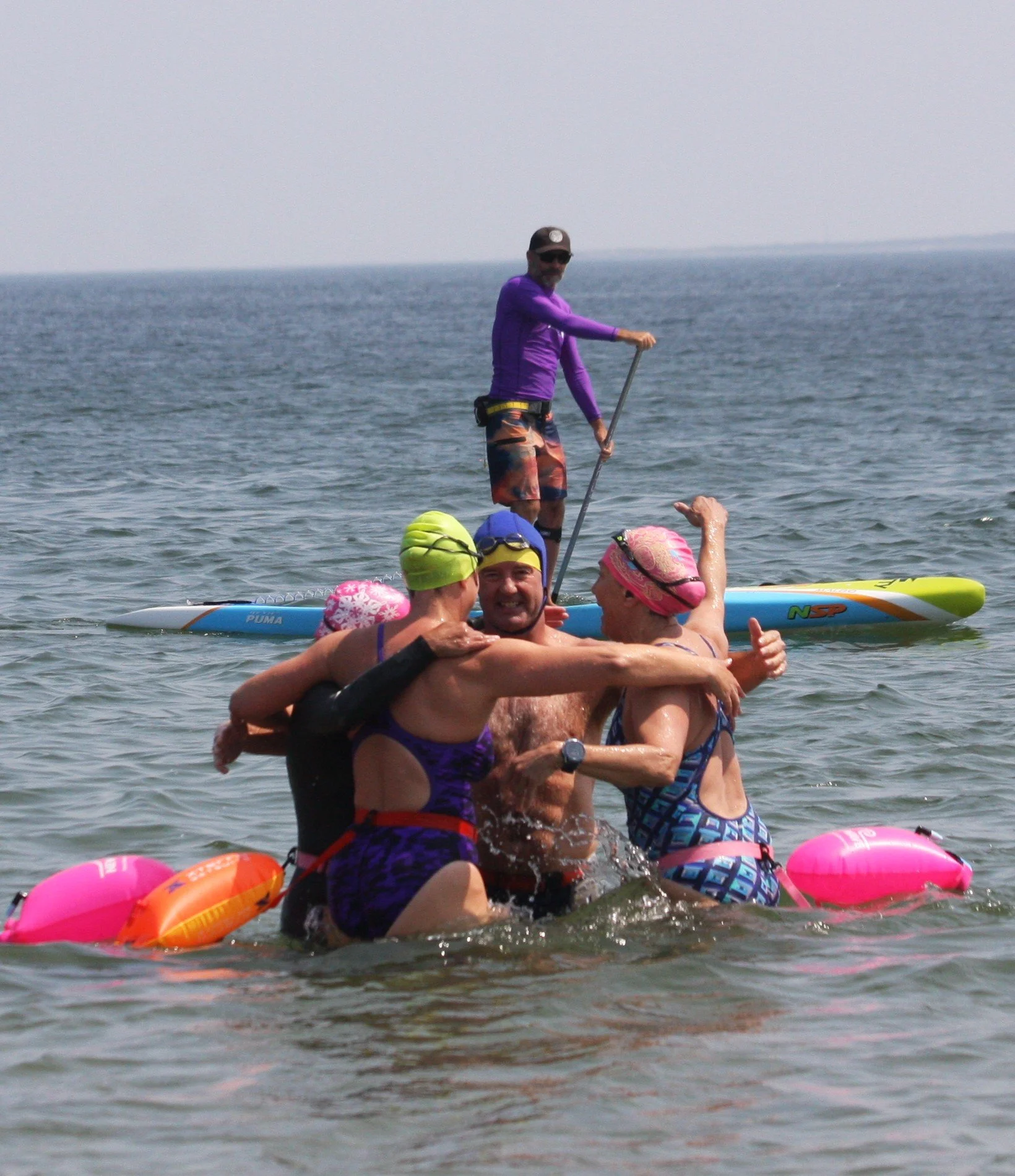 A group of swimmers hugging with a paddleboarder in the background